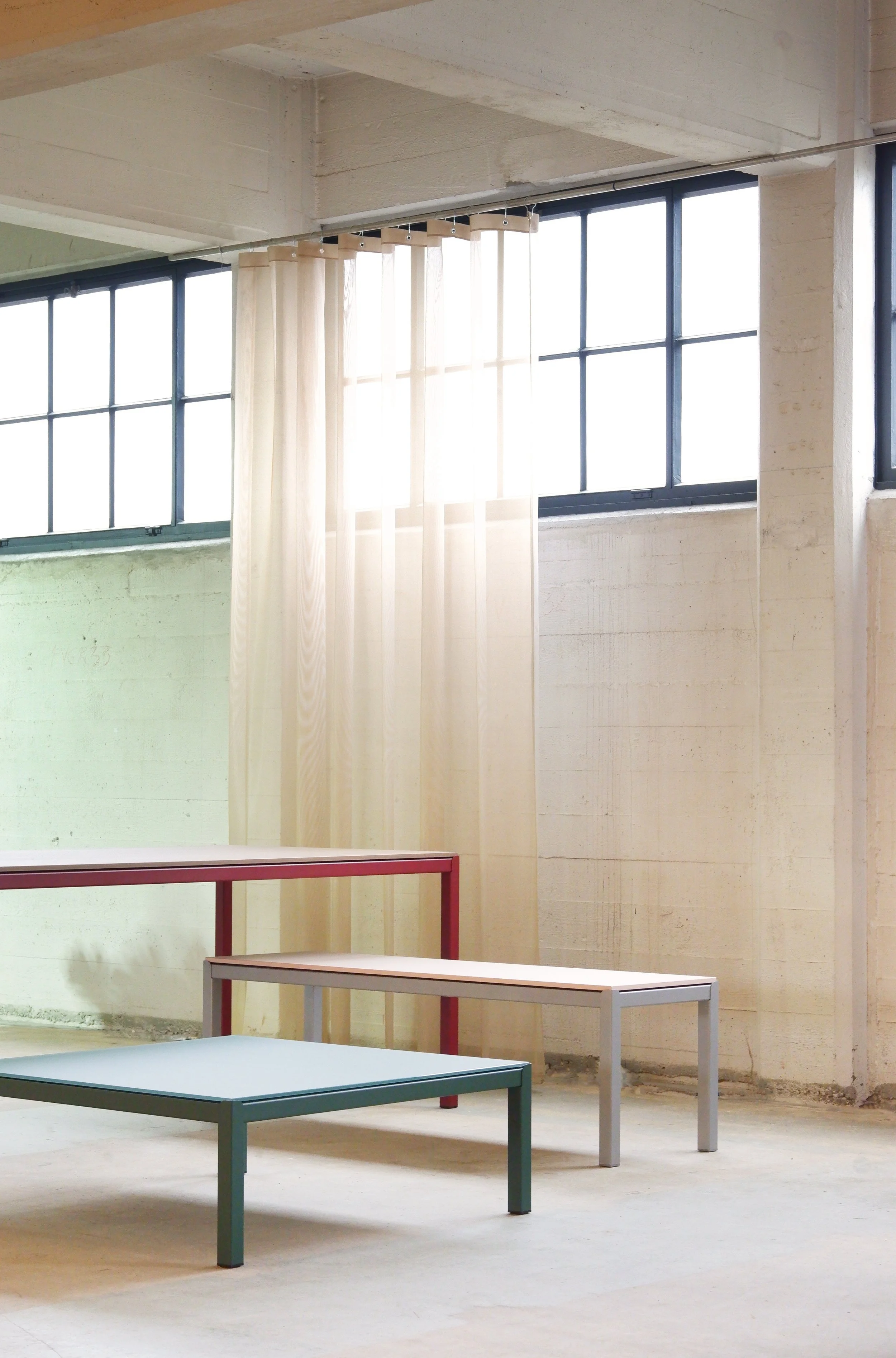 Binnenruimte met grote ramen, doorlaatbaar natuurlijk licht, in een industriële stijl. Interesse in minimalistisch meubelontwerp, met drie verschillende gekleurde tabellen en een lichte gordijn.