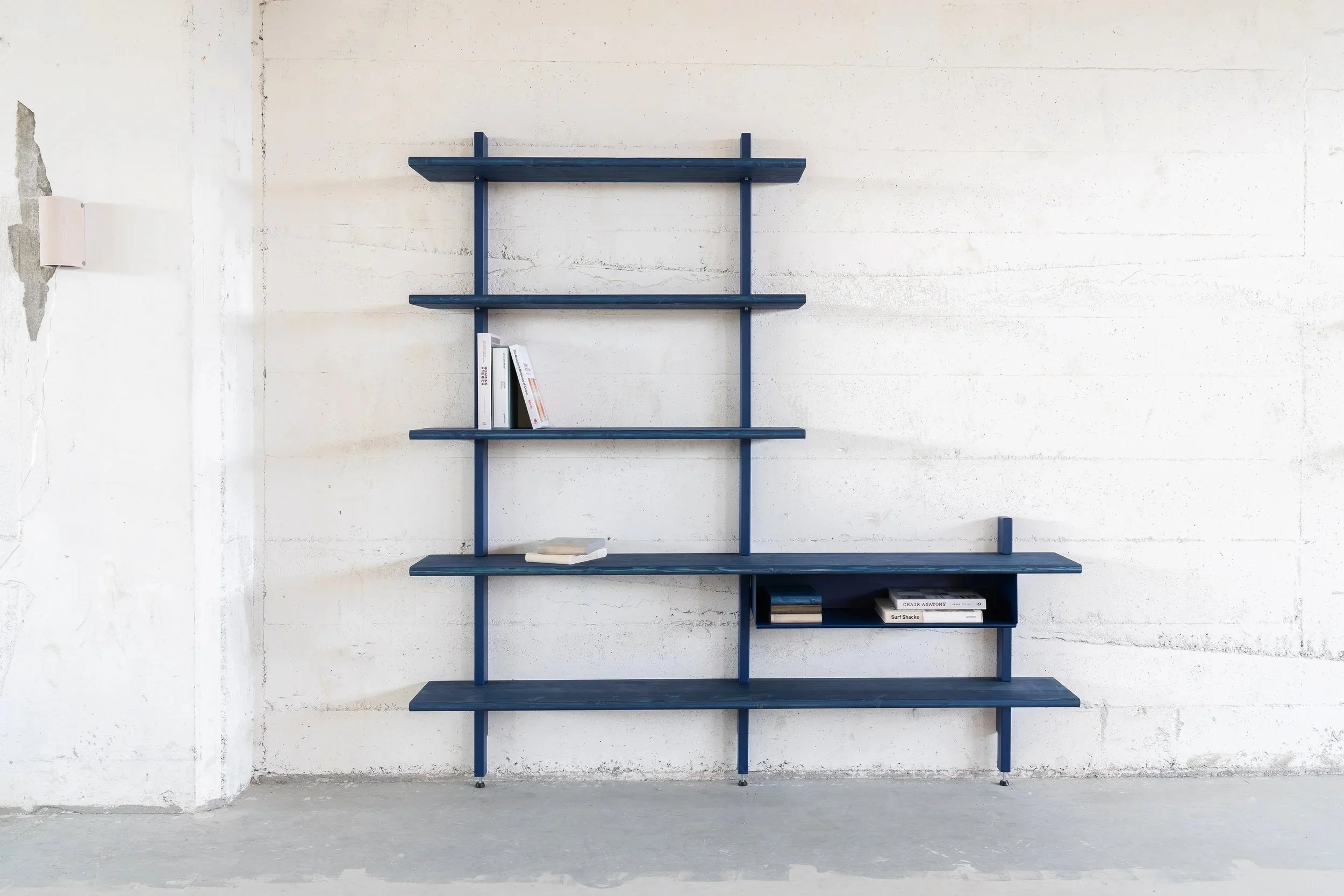 Een muur met een blauw boekenrek met vijf planken, beperkte boeken op sommige planken, tegen een witte betonmuur in een lege ruimte.