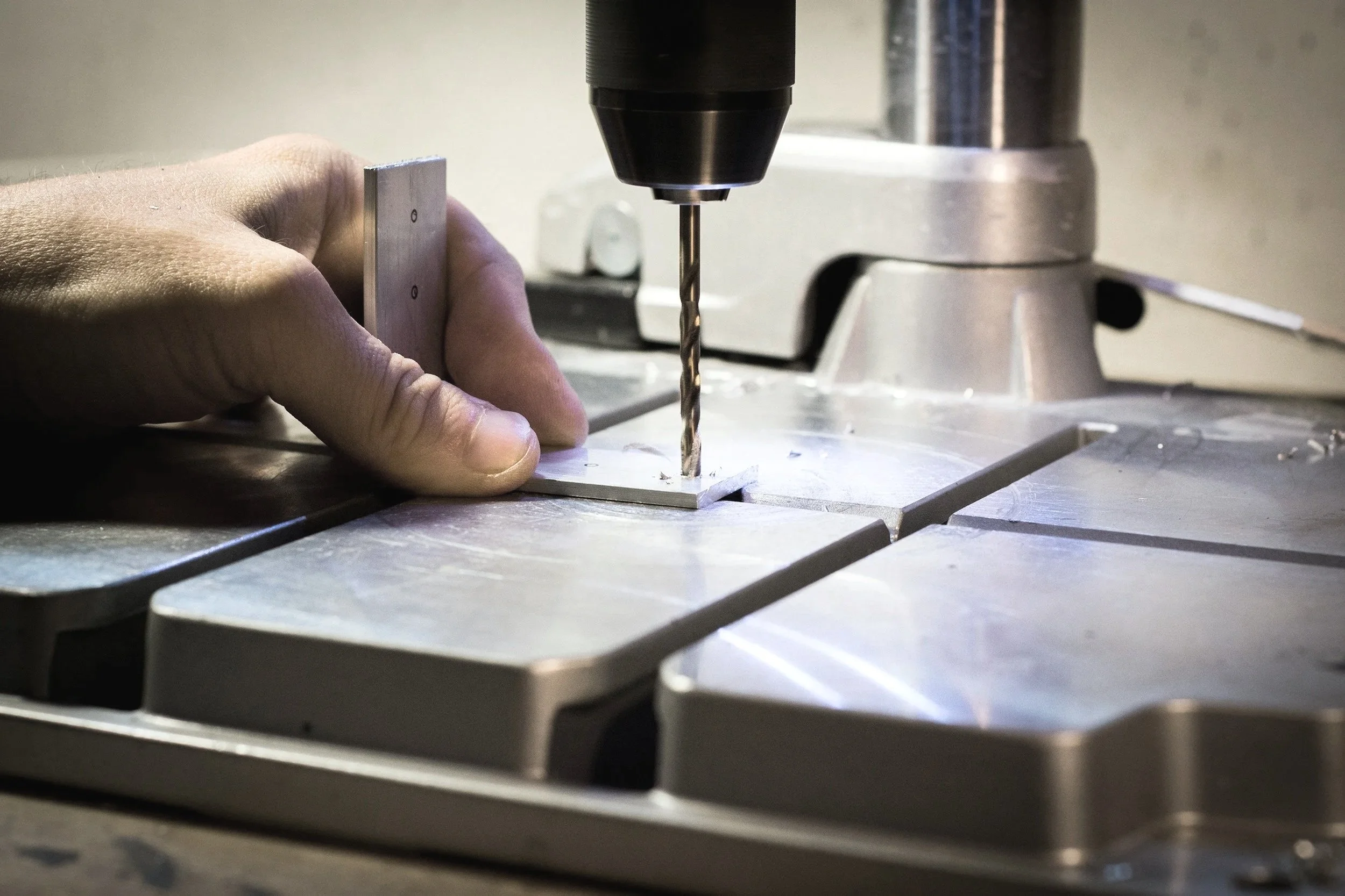 Een hand die een stuk metaal vasthoudt terwijl een boor in een metalen plaat wordt gedraaid.