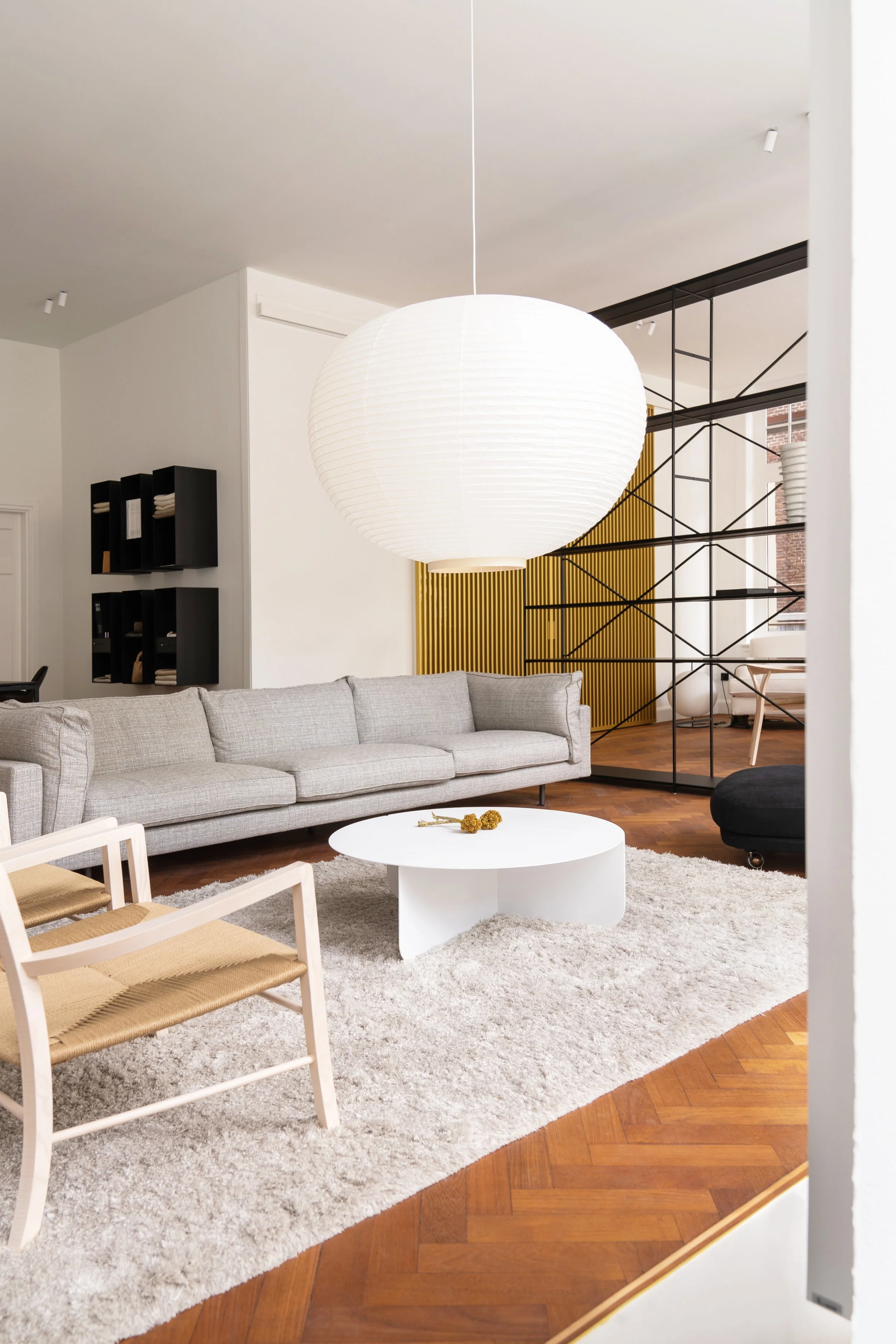 Een stijlvolle woonkamerset met een grote witte bank, een ronde witte salontafel, twee stoelen met houten poten en een witte zitting, een grote witpapieren hanglamp, een zwarte fauteuil, een zachte beige stoffige tapijt, een zwarte glazen boekenkast, een gele decoratieve muur, en een scheidingswand van zwart metalen staven met een witte muur en ramen op de achtergrond.