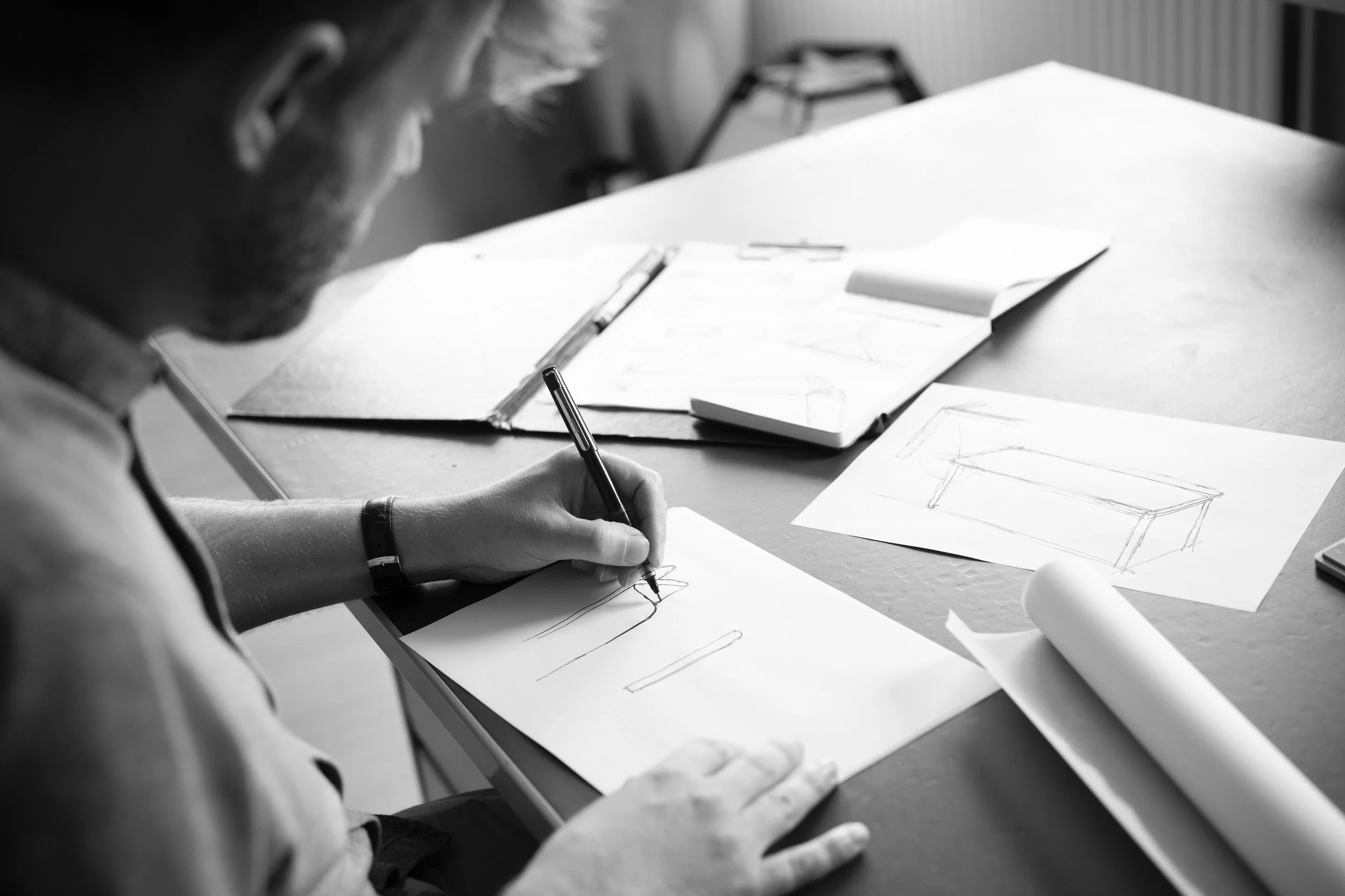 Bas Vellekoop de ontwerper tekent schetsontwerpen op papier met potlood, met meerdere schetsen en tekeningen op tafel. Grijsbeeld.