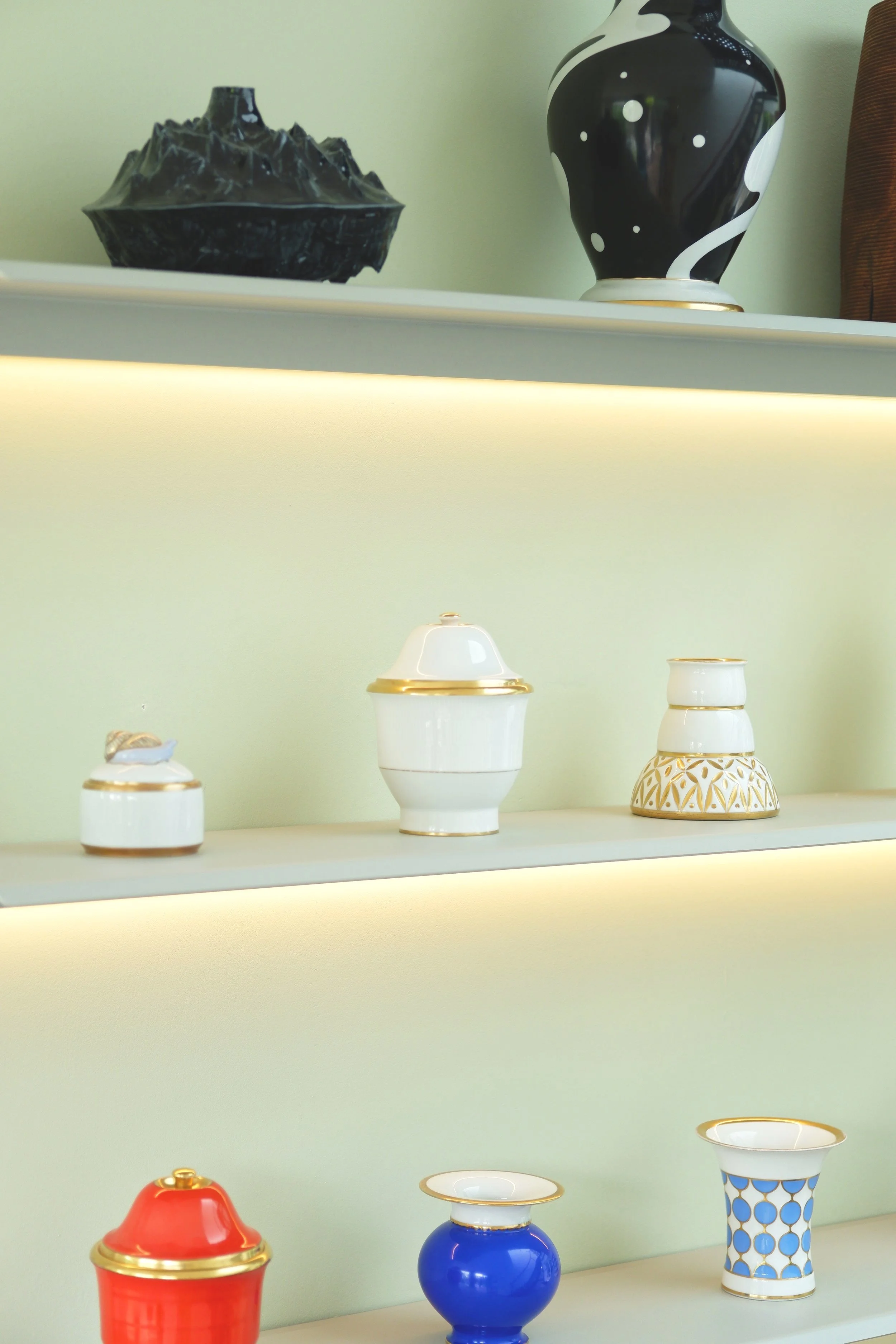 Decoratieve keramische vazen en potten op witte planken met verlichting, diverse vormen en kleuren, waaronder wit, zwart, rood, blauw en goud.