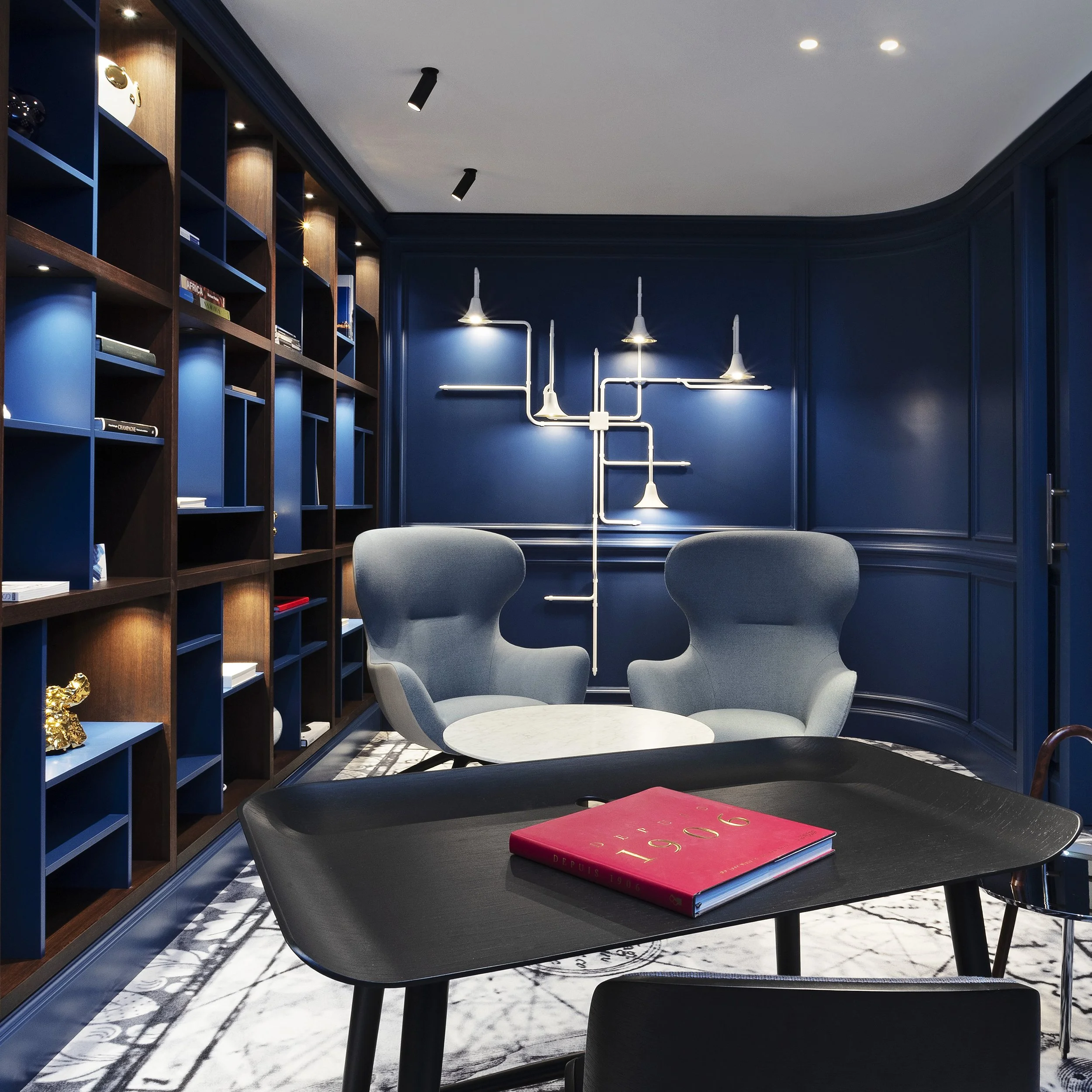 Een moderne lees- of werkkamer met donkerblauwe wanden en een grote houten boekenkast aan de linkerzijde. In het midden twee lichte, comfortabele hoekfauteuils rond een kleine ronde tafel. Aan de muur hangt een decoratief designlicht dat lijkt op een vertakt systeem met meerdere lampjes.