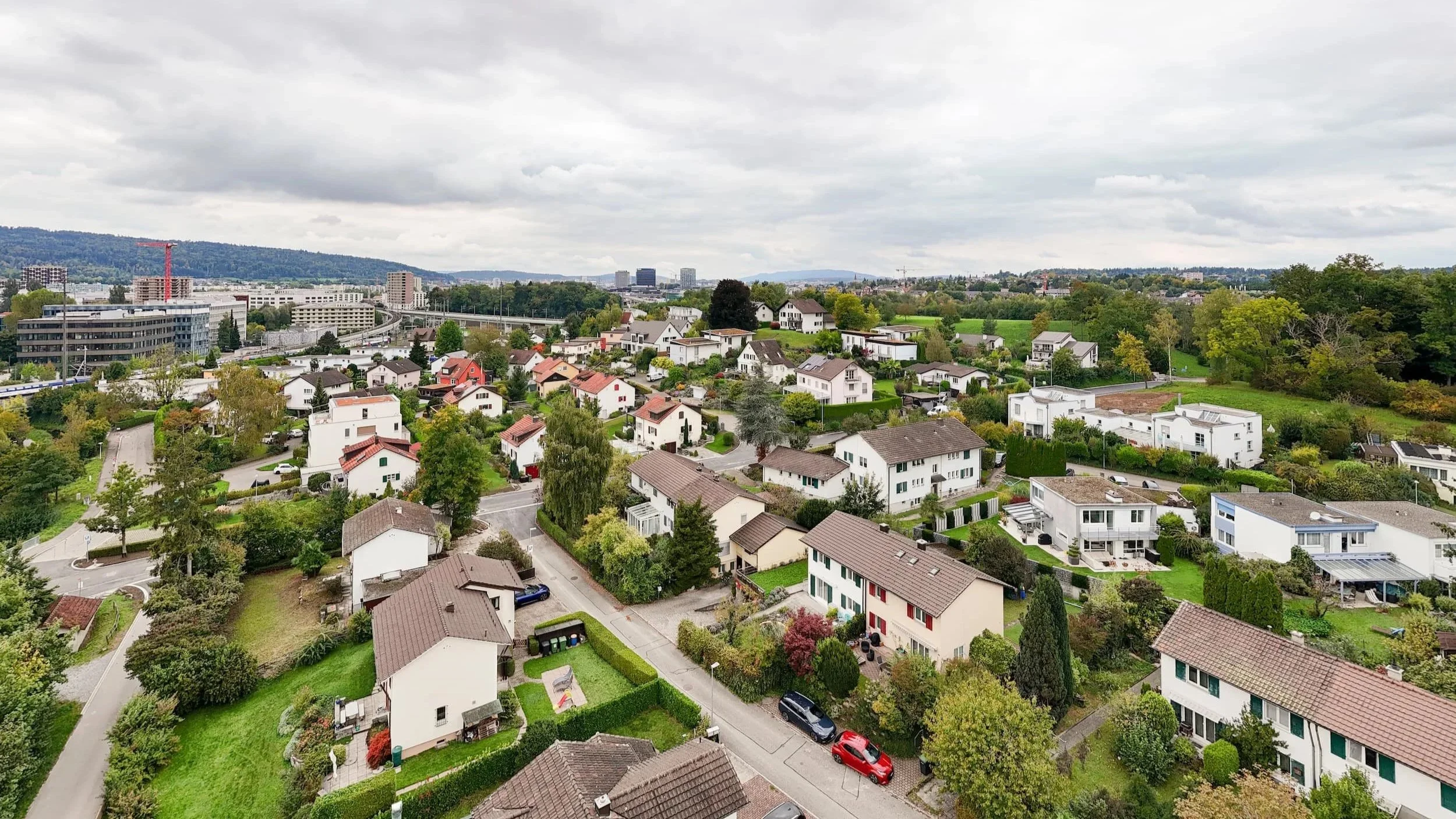 Luftaufnahme eines Wohngebietes mit Einfamilienhäusern, umgeben von Bäumen und Grünflächen, im Hintergrund eine Stadt mit Hochhäusern und grünen Hügeln bei bewölktem Himmel.