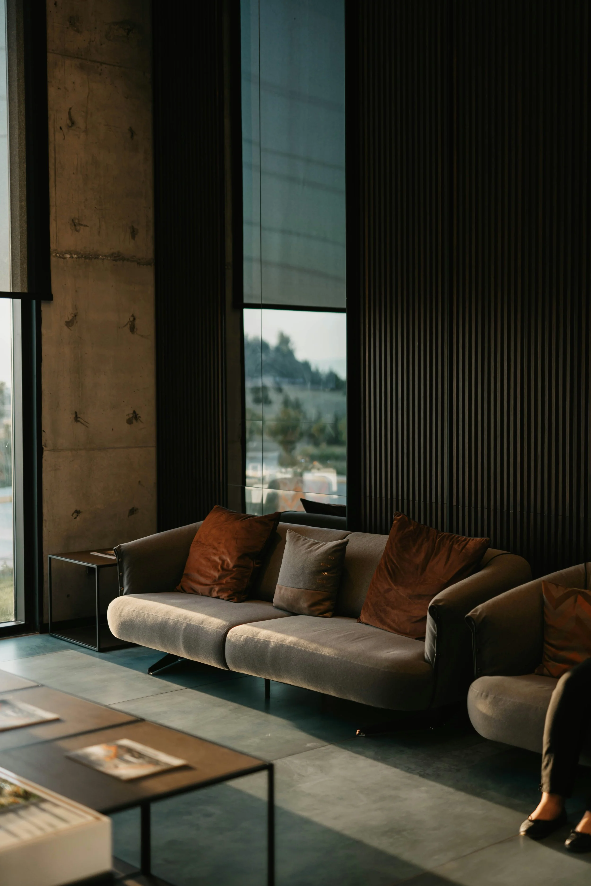 Modern living room with gray sofa decorated with brown and gray pillows, large windows with black blinds, and a coffee table with magazines.