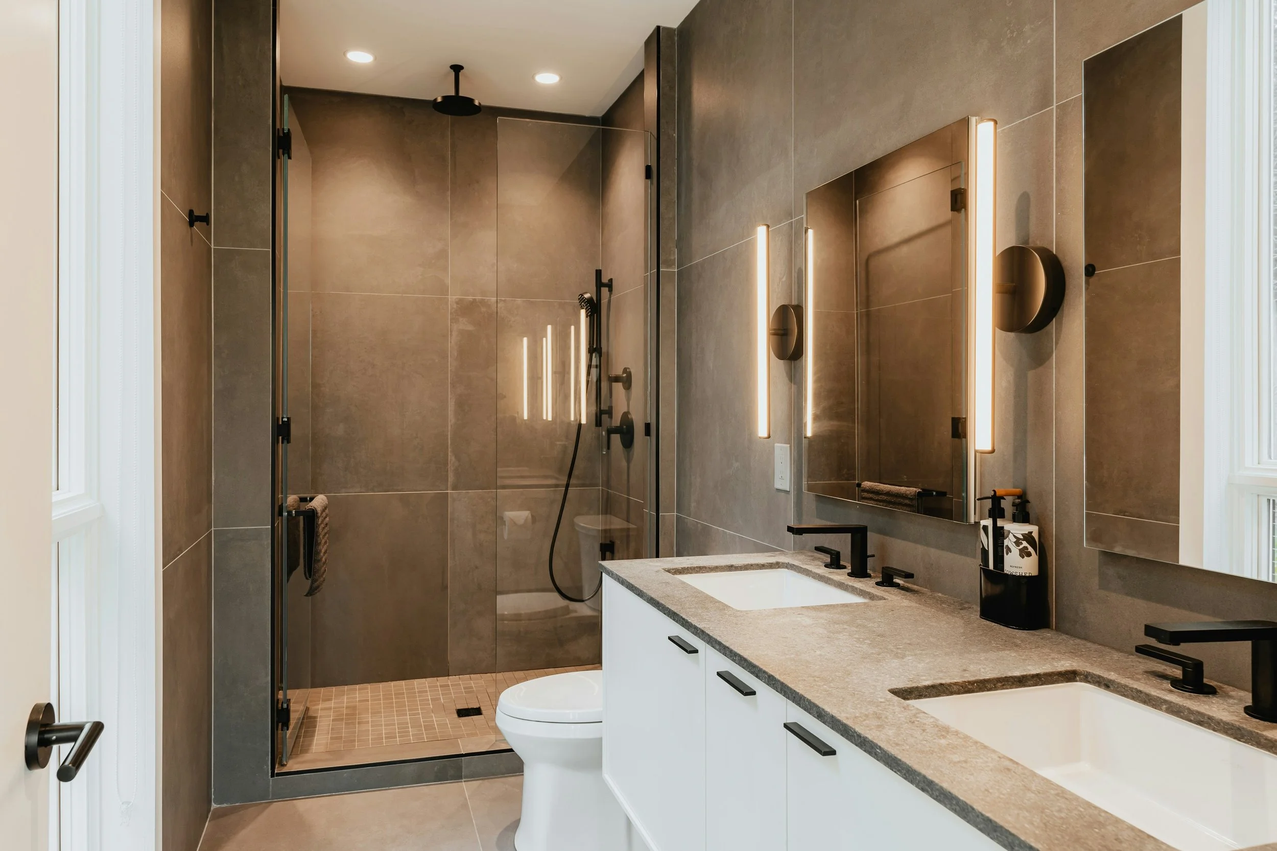 Modern bathroom with dual sinks, black fixtures, large mirrors with vertical lighting, and a walk-in shower with black hardware and beige tiles.