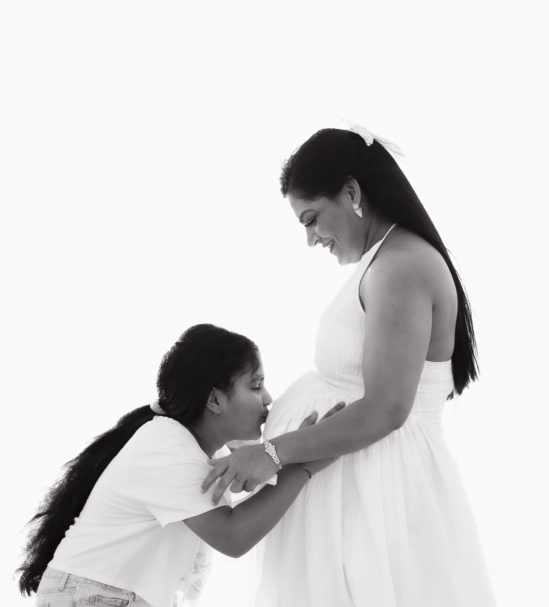 A woman in a white dress pregnant with a young girl kissing her baby bump.