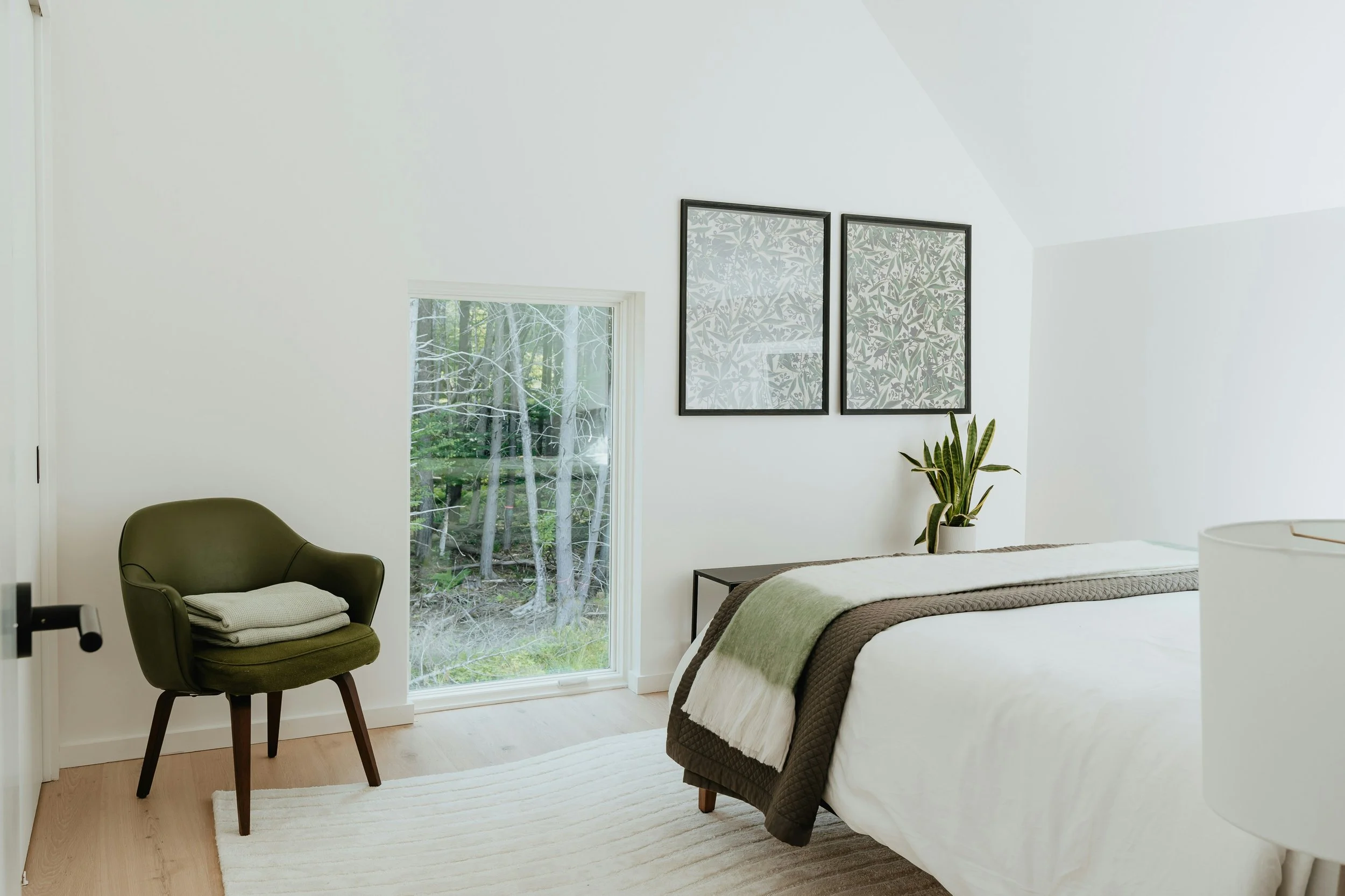 A minimalist bedroom with white walls, a bed with white and green bedding, a small black side table with a potted plant, two framed botanical prints, a green armchair with folded blankets, and a large window showing a view of trees outside.
