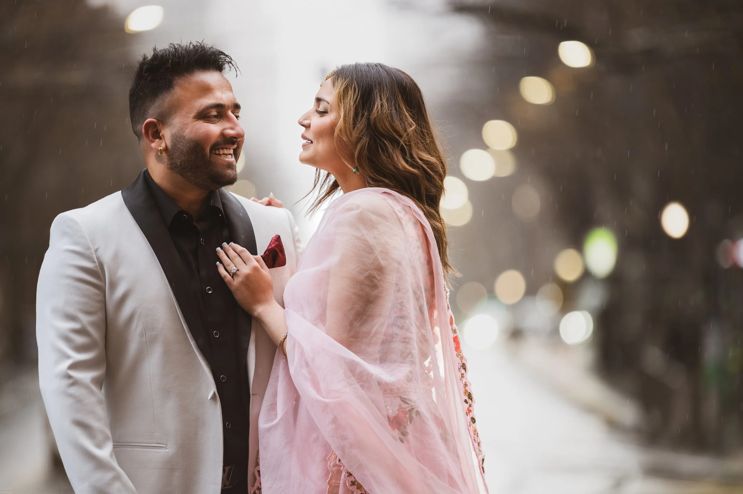A joyful man and woman in formal attire smiling at each other under a rainy sky, standing close together on a street with blurred bokeh lights in the background.