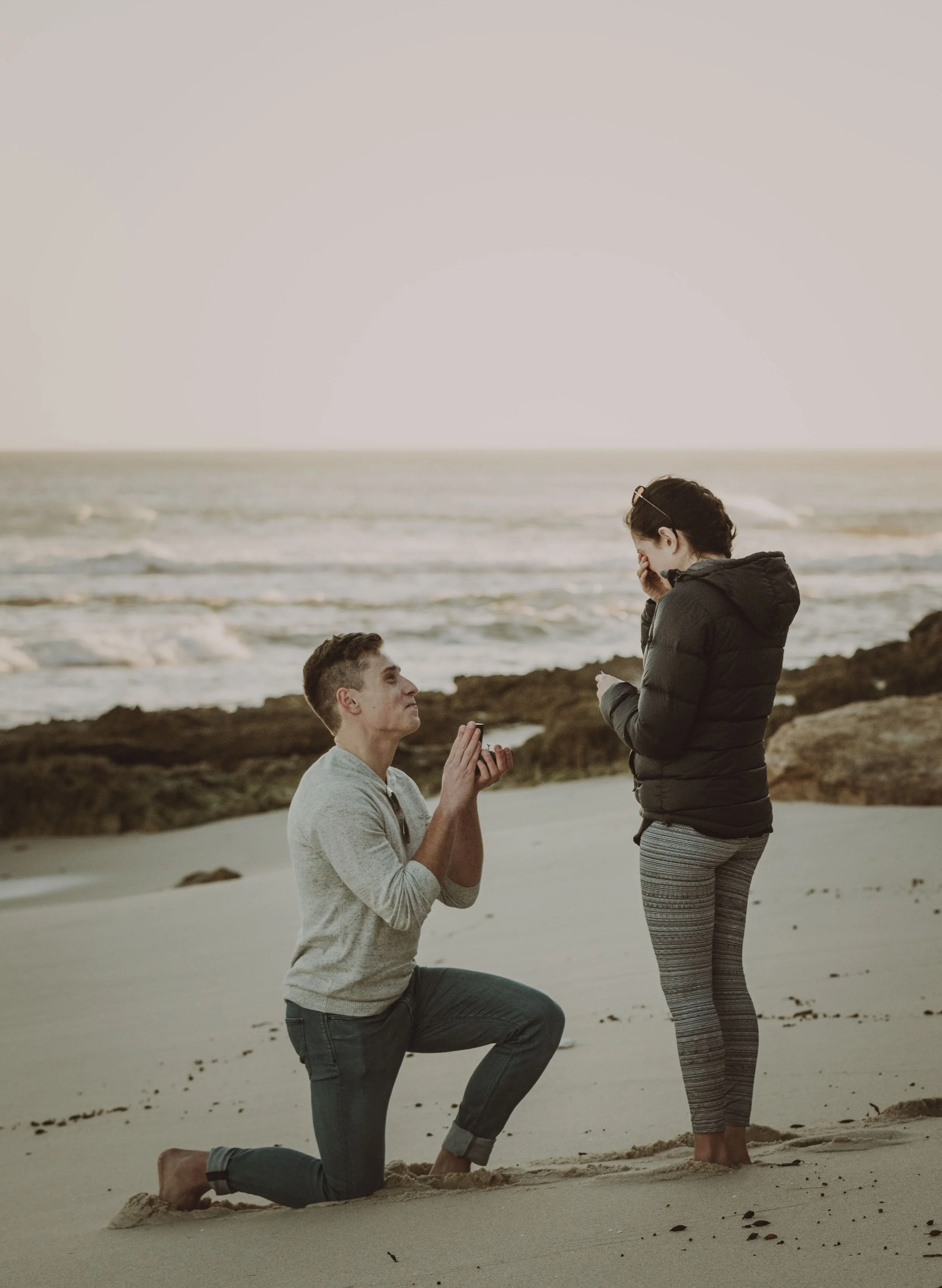 A man on one knee proposing to a woman on a sandy beach, with rocks and ocean waves in the background during sunset.