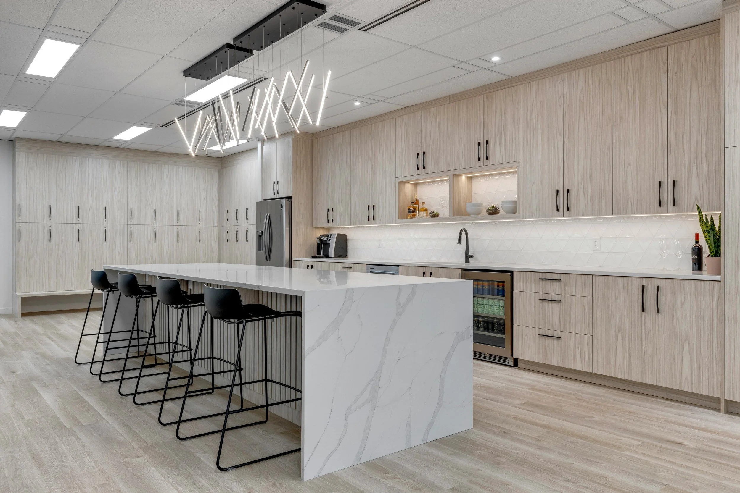 Modern kitchen with light wood cabinets, a marble island, black chairs, and minimalist lighting fixtures.