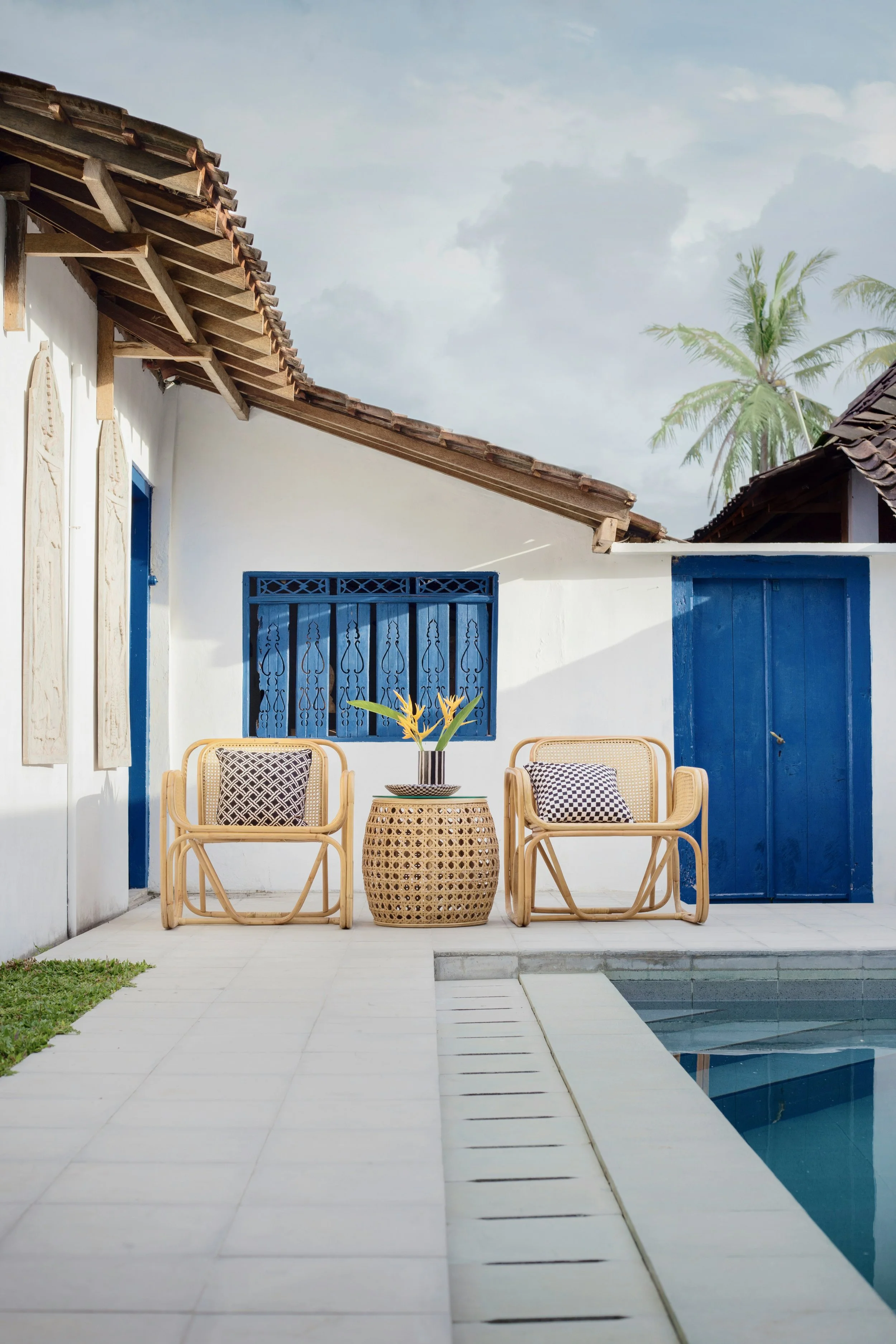 Two rattan chairs with patterned cushions beside a woven side table with a potted plant, set against a white wall with blue window and door, near a swimming pool, with palm trees in the background.
