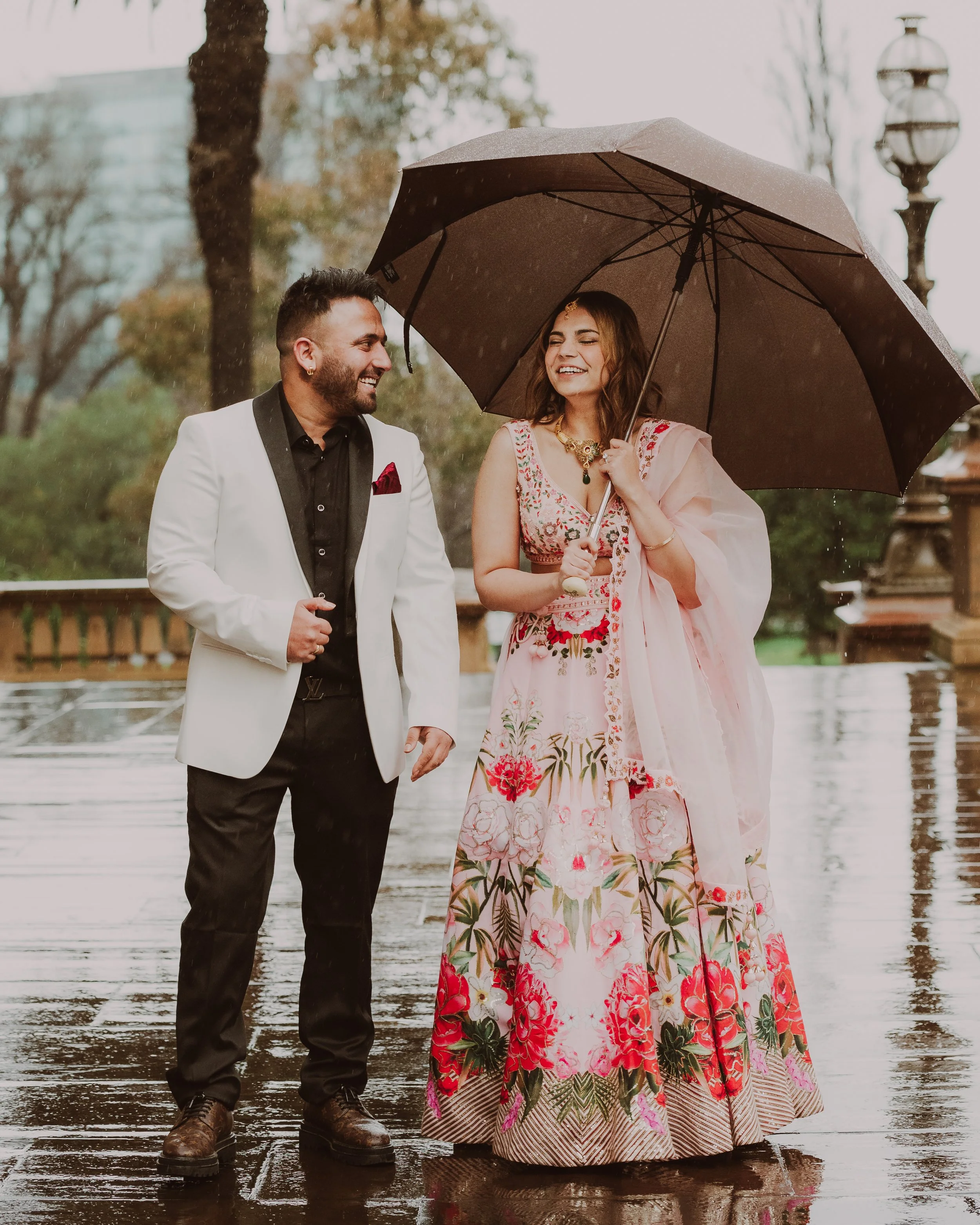 A man and woman standing together outdoors in the rain, smiling, with the woman holding an umbrella over them. The woman is dressed in a pink floral traditional dress, and the man is wearing a white suit jacket with a black shirt and pants.