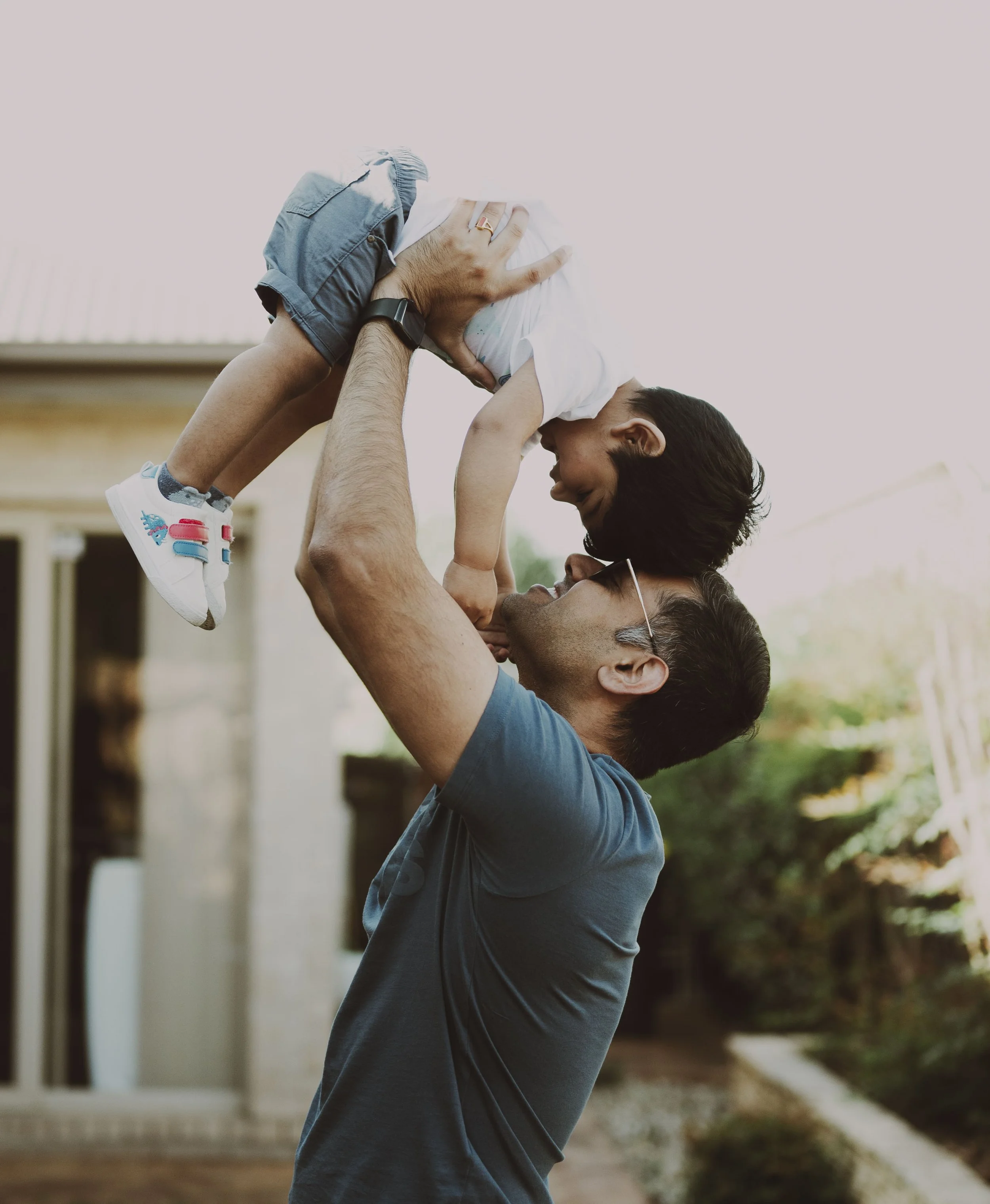 An adult male lifts a young boy into the air outdoors, both smiling and touching noses, in a backyard setting.