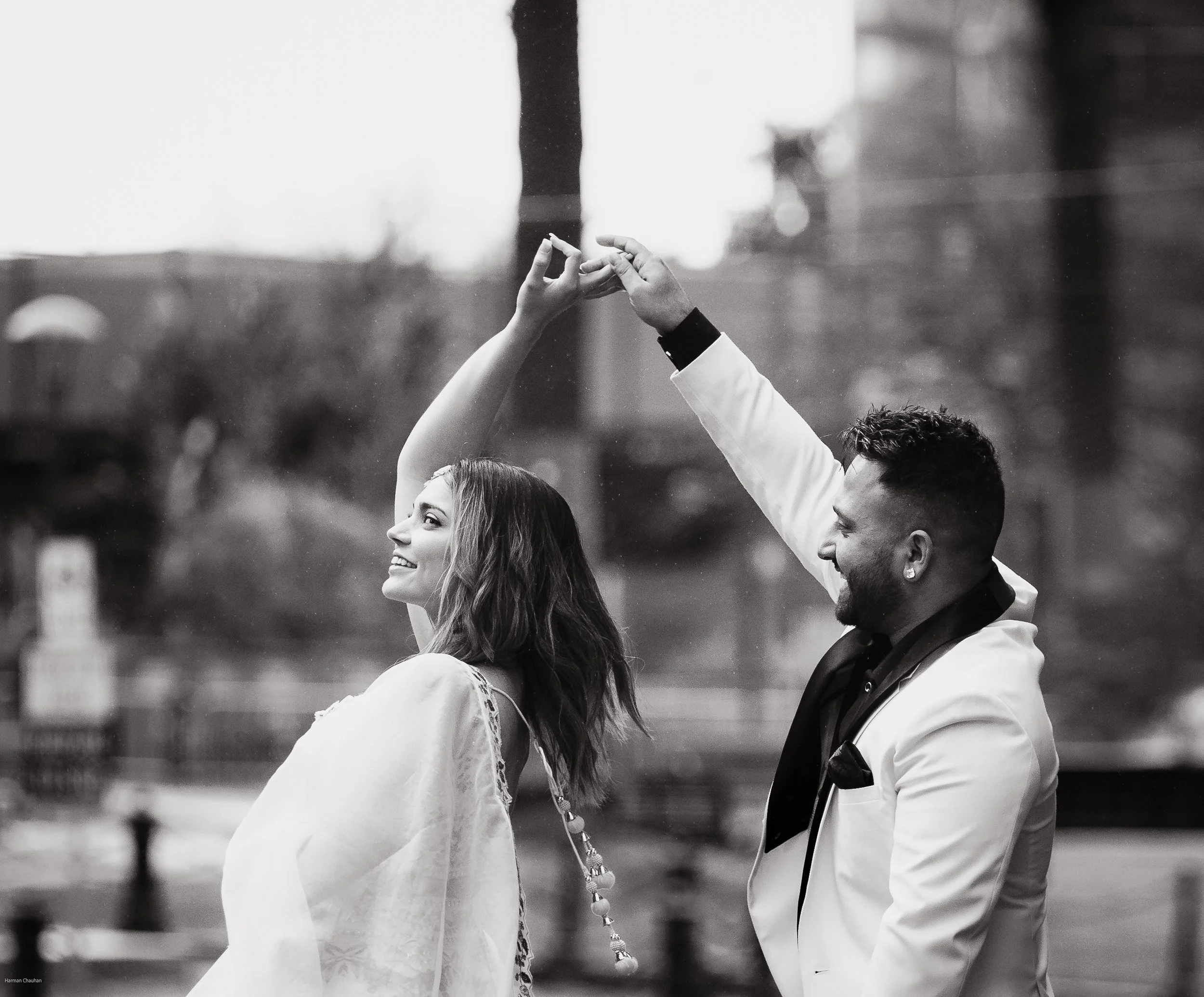 A black and white photo of a happy couple dancing outdoors, with the woman smiling and the man wearing a white tuxedo jacket, both holding hands above their heads.