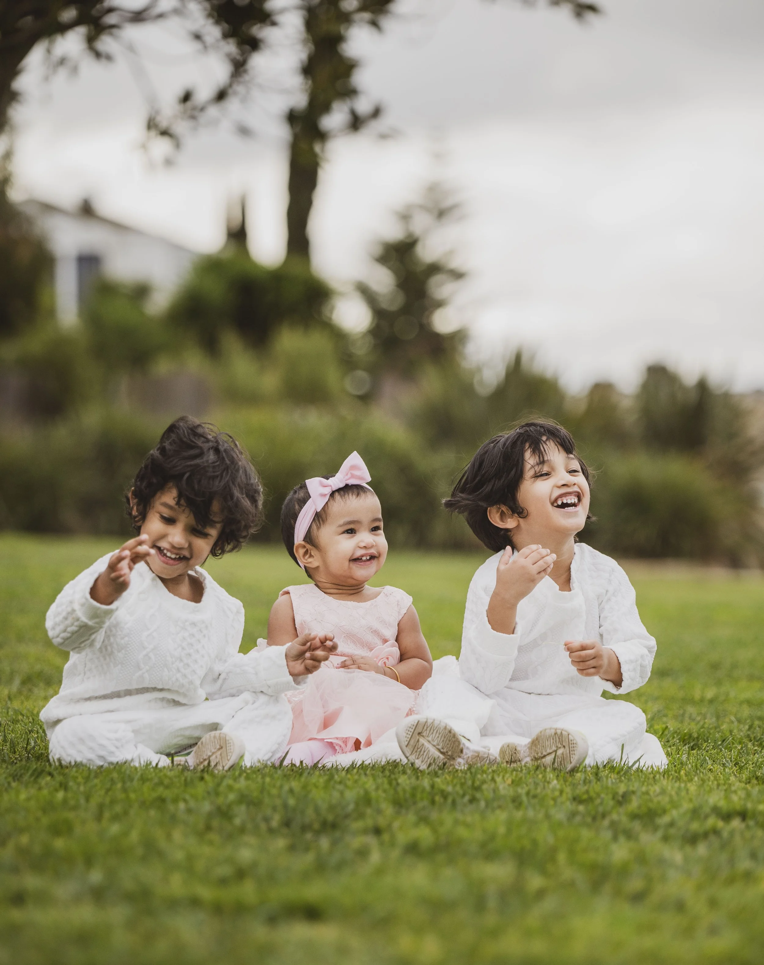Three young children sitting on grass outside, smiling and laughing, with a blurred background of trees and sky.