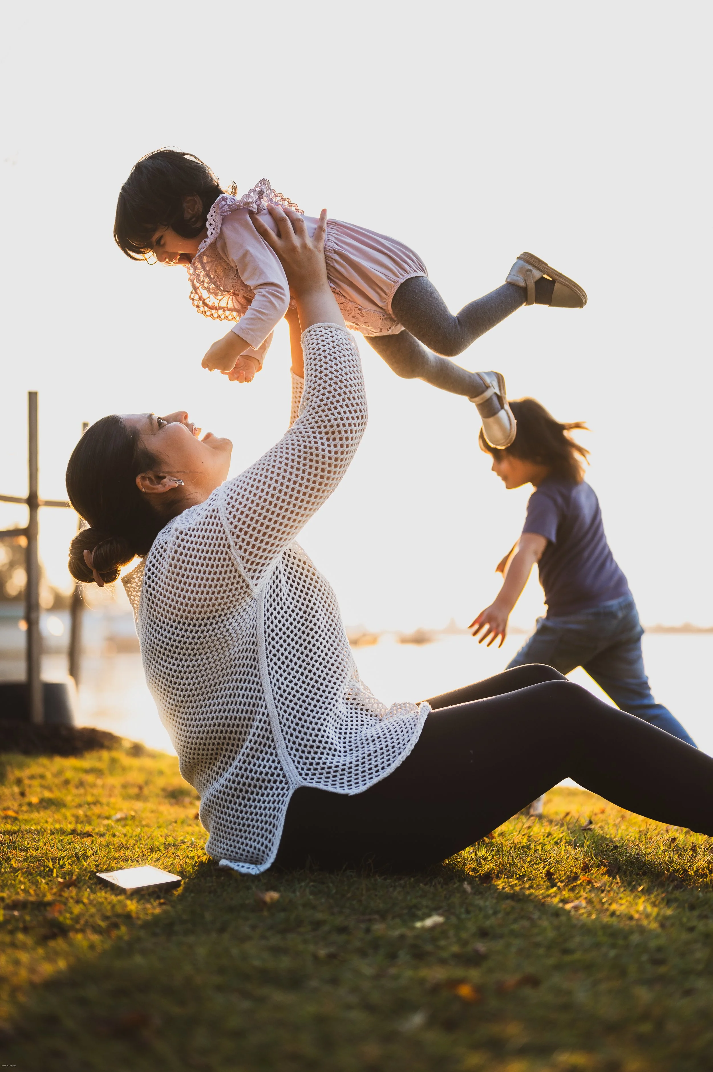 A woman sitting on the grass lifting a young girl in the air, with another girl playing in the background near a body of water during sunset.