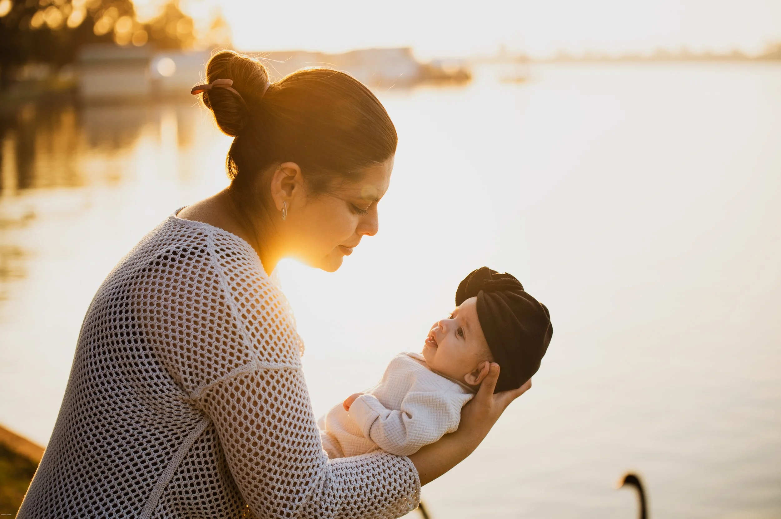 A woman holding a baby by a lakeside during sunset, with ducks swimming nearby.