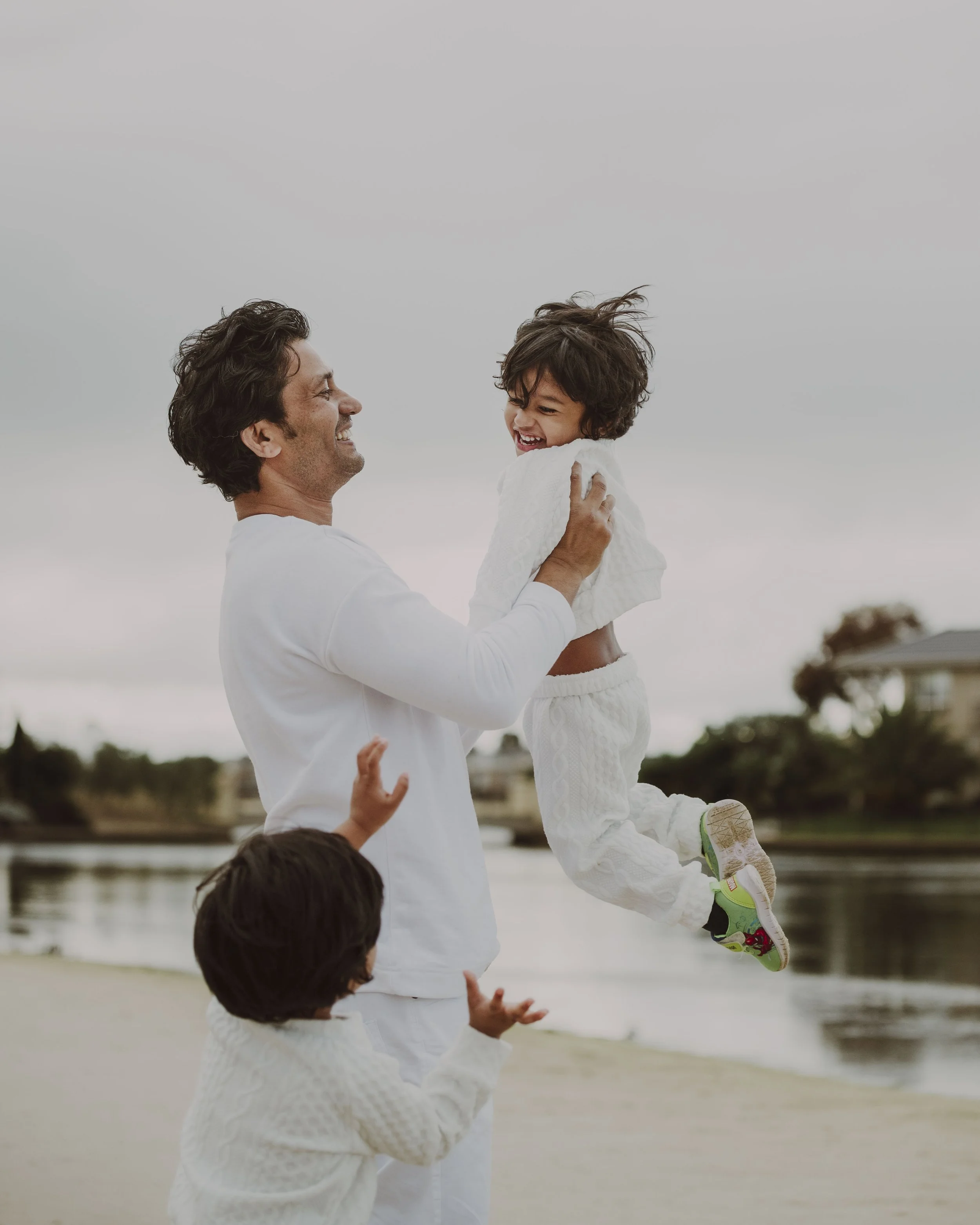 A man playing with two children by the water on a cloudy day, with one child in the man's hands laughing and the other reaching up.