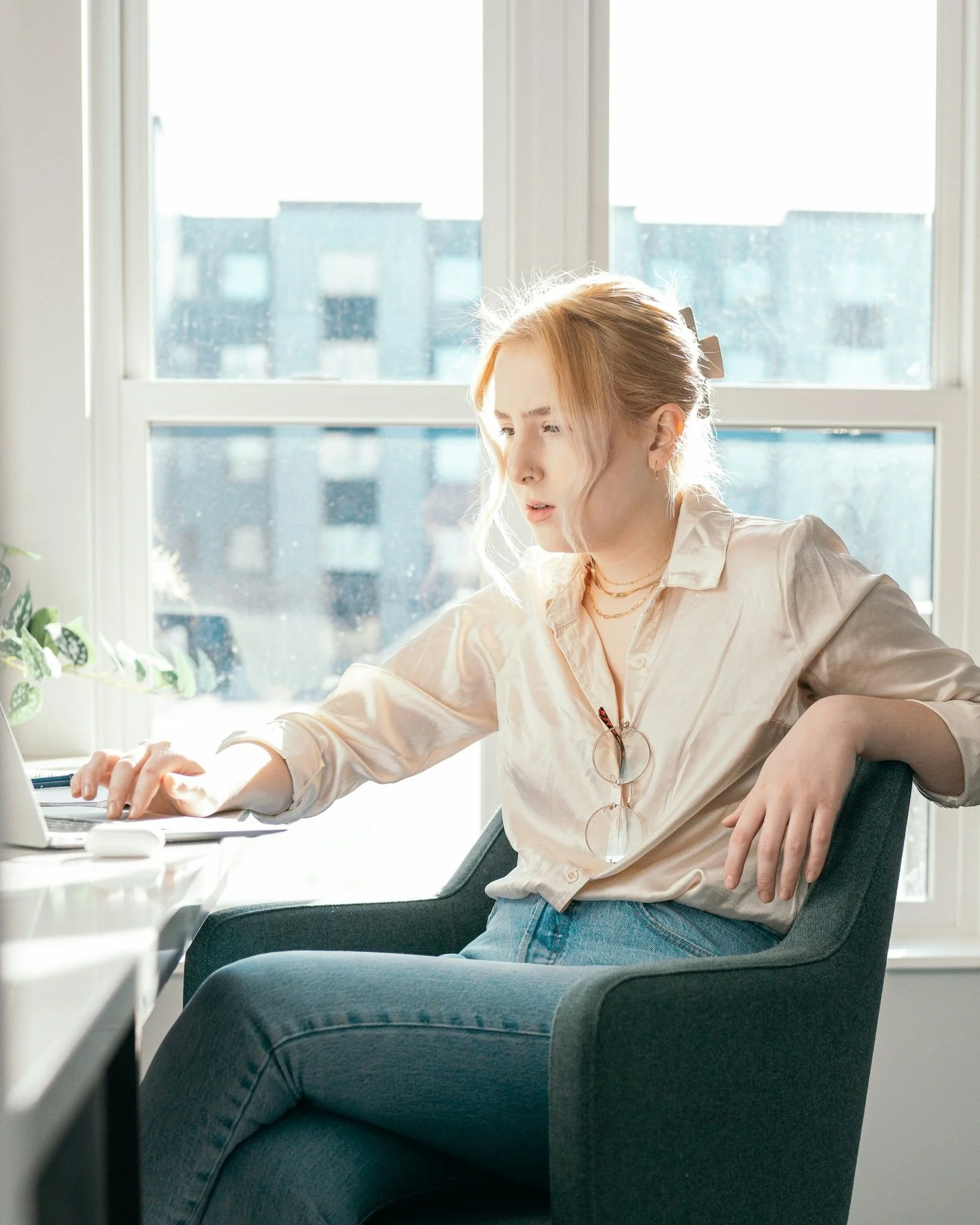 A young woman with blonde hair seated near a large window, working on a laptop on a desk in bright sunlight.