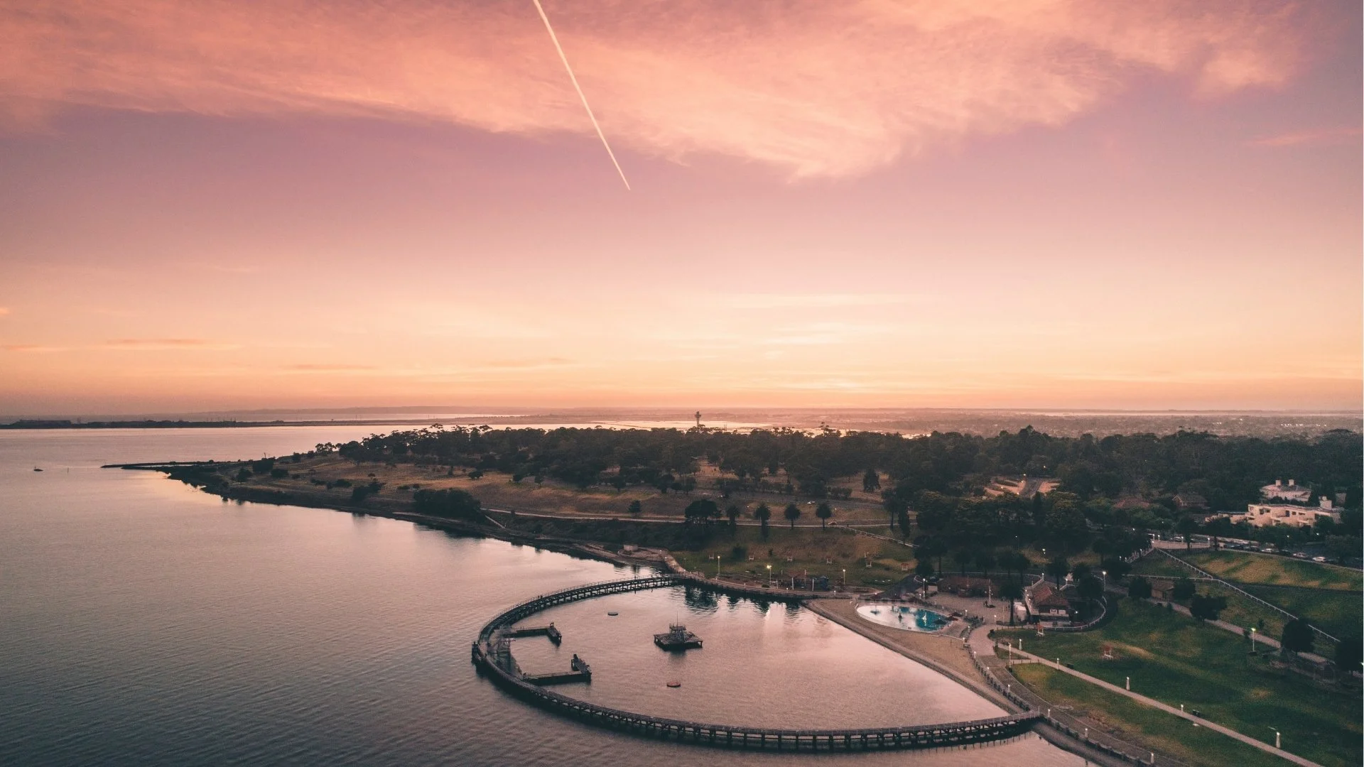 Aerial view of the Geelong waterfront, including Eastern Beach boardwalk during sunset, with trees, houses, and a park in the background, under a pink and orange sky.
