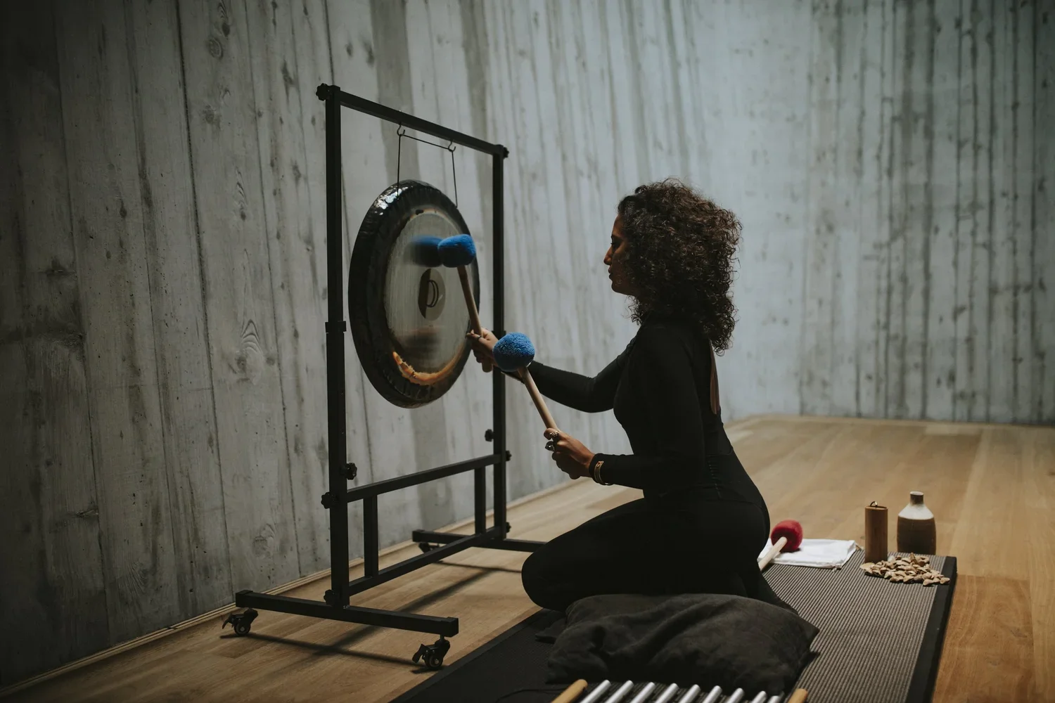 A woman with curly hair kneeling on a yoga mat, playing a gong with a mallet. She is in a room with wooden floors and a wood-paneled wall, and various percussion instruments and objects are nearby.