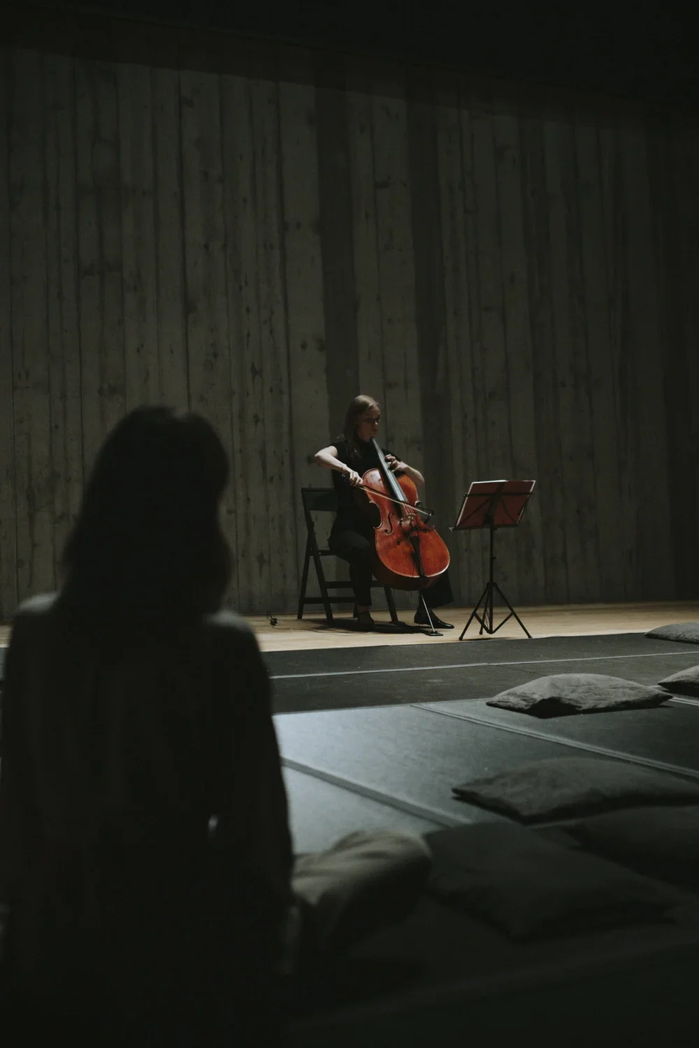 A woman playing cello on stage with music stand, in a dimly lit room with wooden wall background, audience member seated in the foreground.