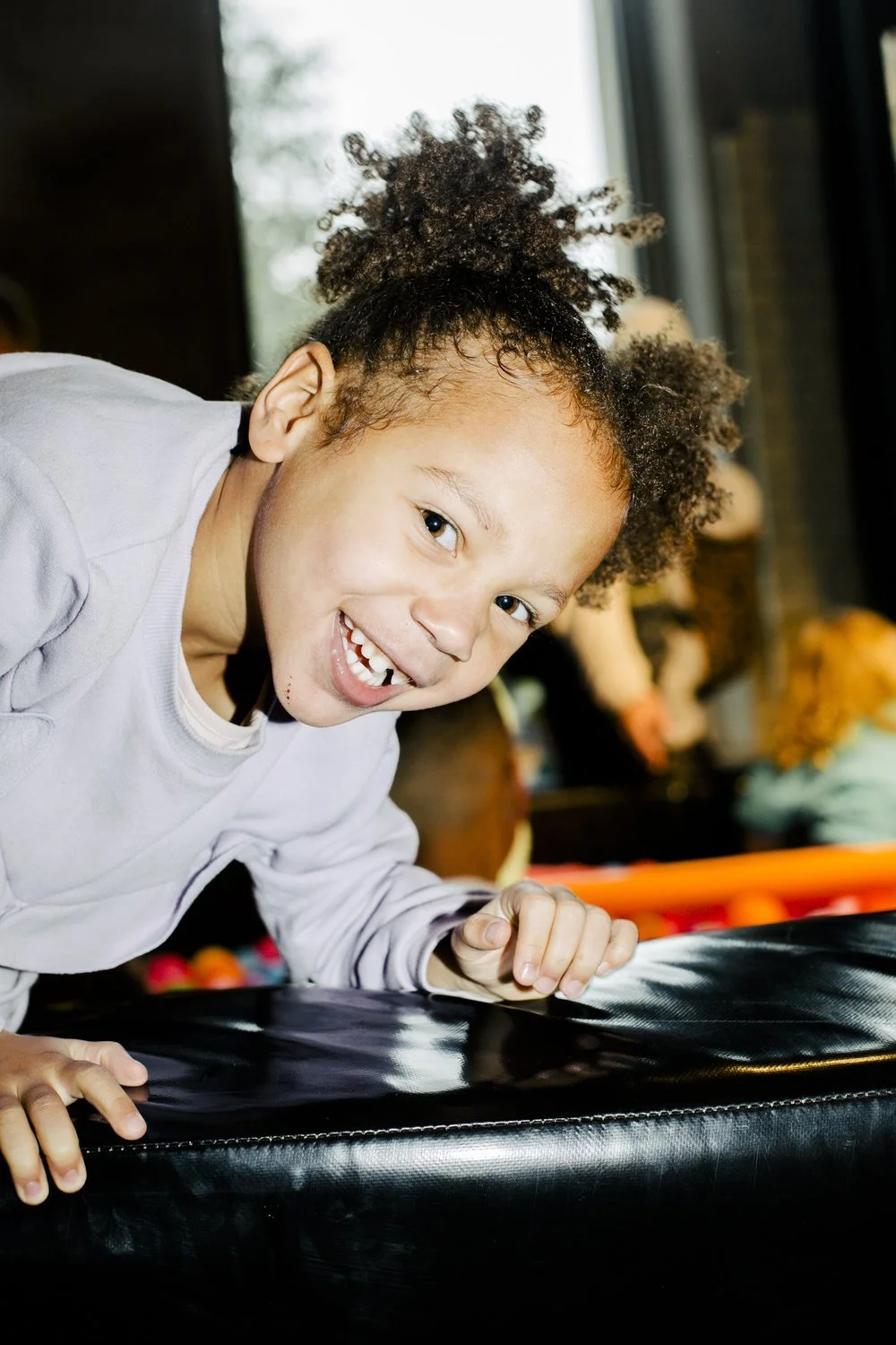 Young girl smiling and leaning over a trampoline with black padding, in an indoor or outdoor play area with blurred background.