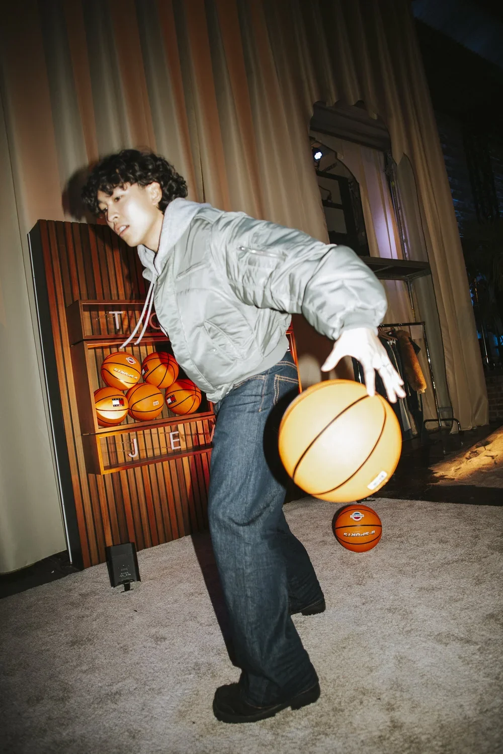 A young man with curly hair wearing a grey bomber jacket and jeans is playing with a basketball indoors. He appears to be in motion, possibly spinning or dribbling the basketball, with a wall of basketballs and curtains in the background.