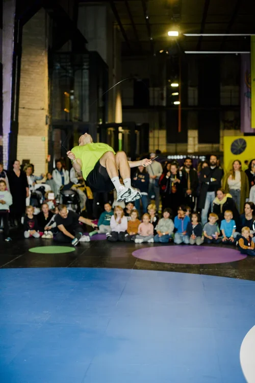 A man in a bright green shirt and black shorts is mid-air performing a jump on a slackline at an indoor event, with an audience of children and adults watching from matted seating.