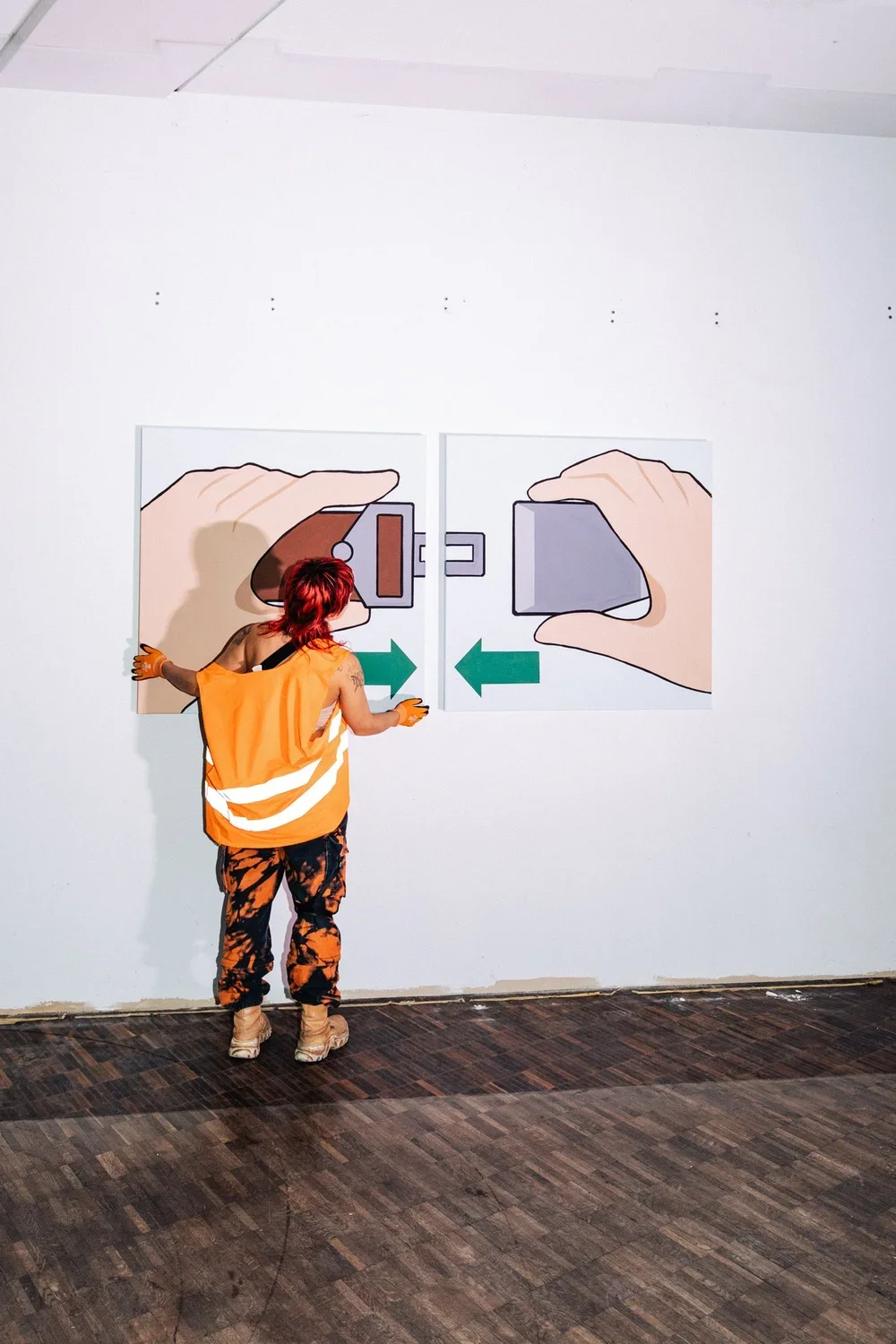 Person with red hair wearing an orange safety vest and camouflage pants interacting with a large wall graphic showing two hands holding a key and a padlock with arrows pointing in both directions.