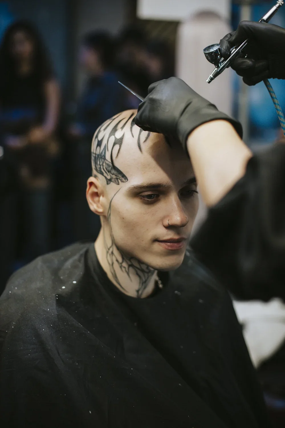 A person with a shaved head getting a black tattoo on the scalp, with tattoo artist wearing gloves using tattooing equipment, while in the background several people are observing.