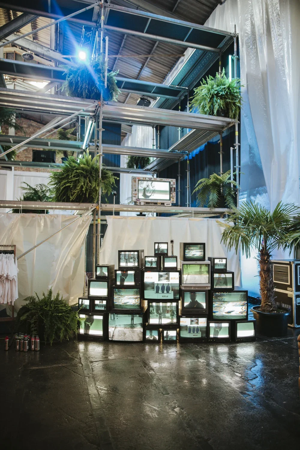 An art installation featuring stacked vintage CRT televisions displaying static and distorted images, surrounded by lush green potted plants, in an industrial-style space with metal scaffolding and a concrete floor.