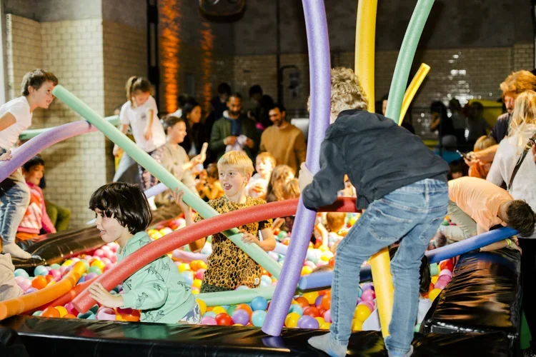 Children playing in a ball pit at an indoor amusement center, with colorful inflatable tubes and adults supervising.