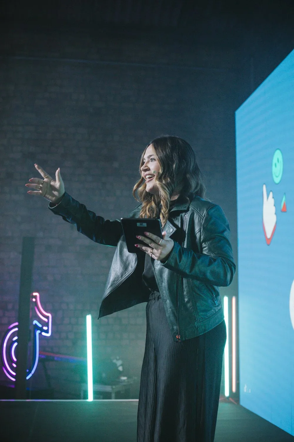 A woman in a leather jacket speaking or presenting with a digital screen behind her showing colorful icons in a modern indoor setting.