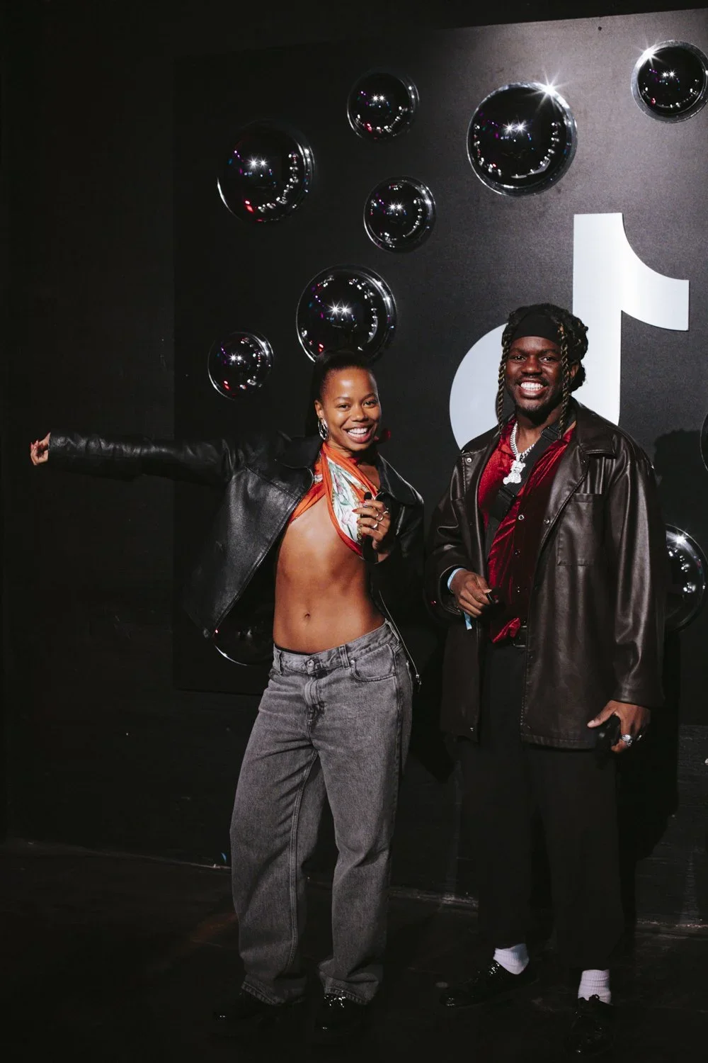 Two smiling individuals standing in front of a black wall with reflective black spheres and a white logo; the woman on the left is wearing a leather jacket, open at the front, with no shirt underneath, and gray jeans, showcasing her midriff; the man 