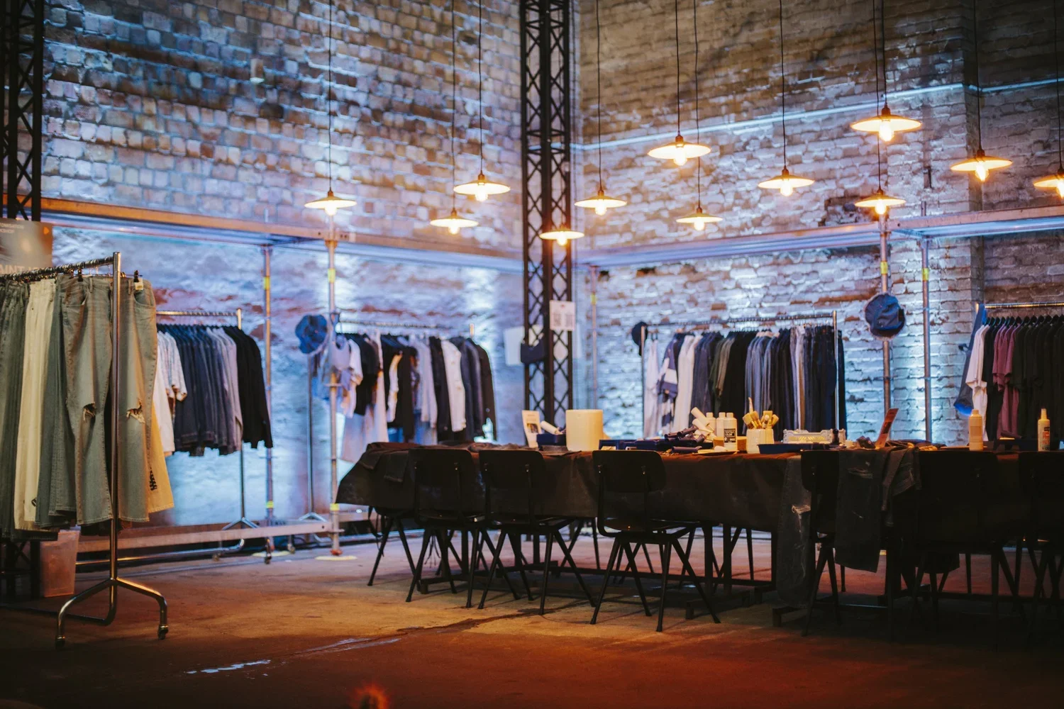 Indoor clothing store with exposed brick walls, hanging lights, clothing racks with shirts and pants, black chairs around a table with supplies, and a casual industrial atmosphere.