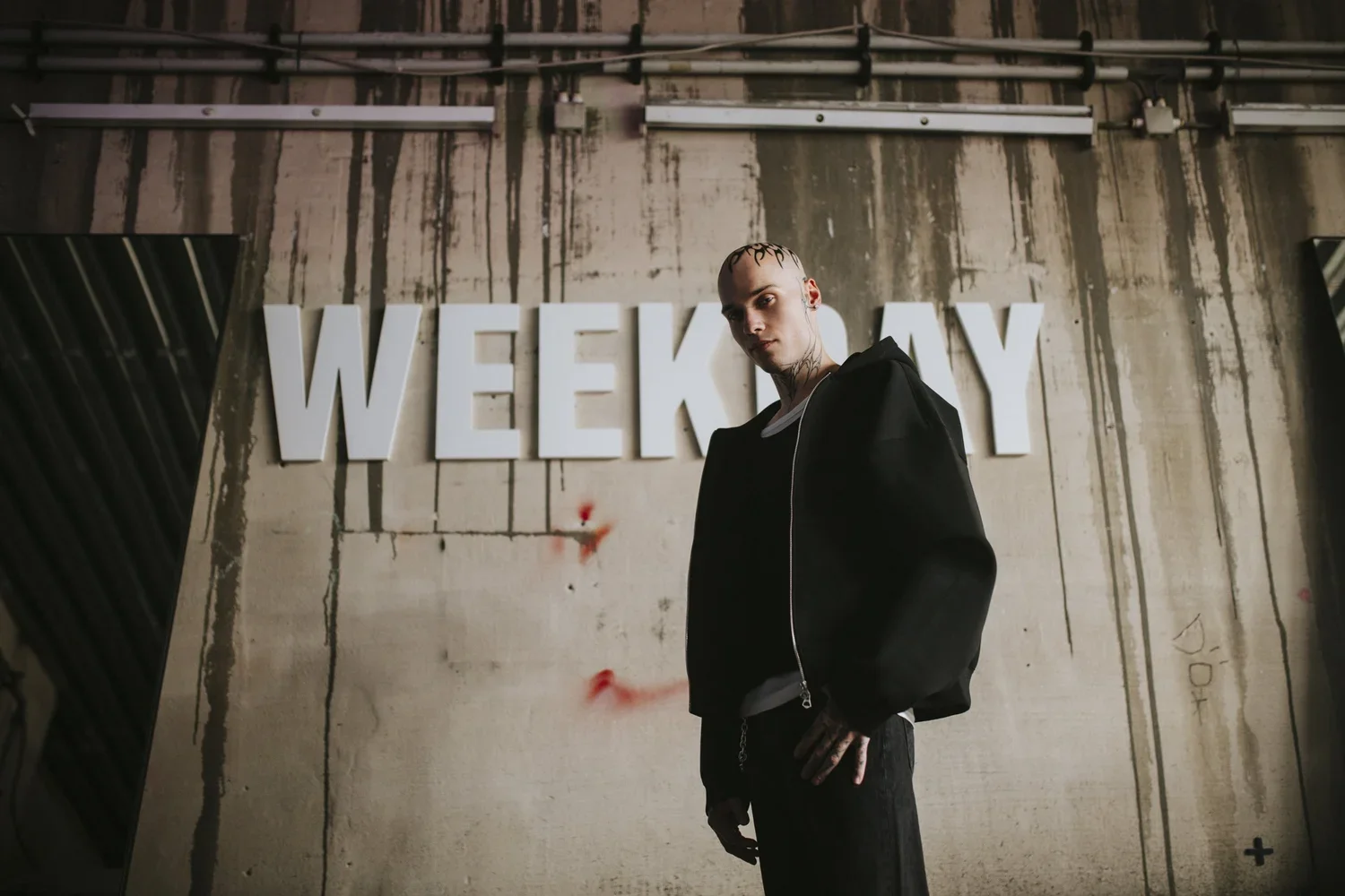 A young man with shaved head and tattoos standing with one hand on his hip in front of a wall with large white letters spelling "WEEKLY."