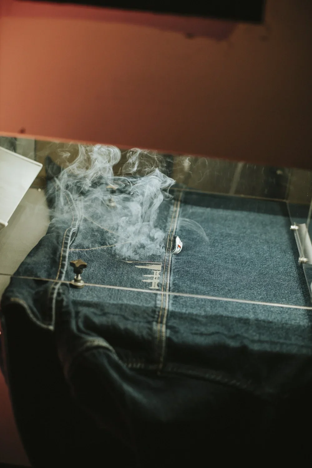 Pair of blue jeans with steam or smoke rising from them, viewed through glass behind a red surface.