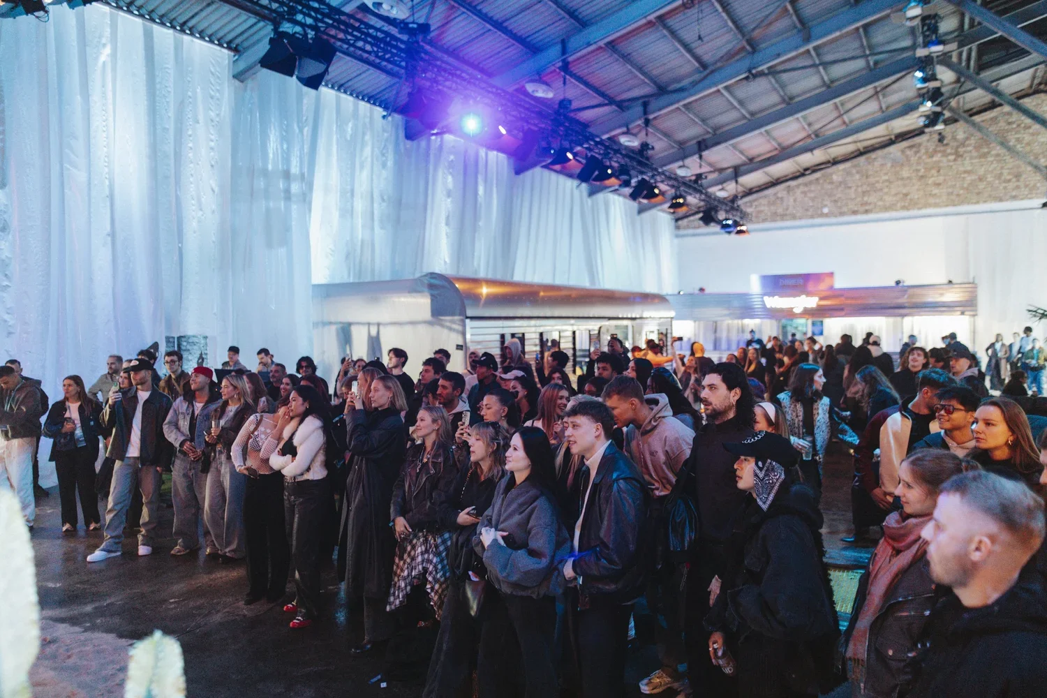 Crowd of people at an indoor event with blue and purple lighting, white draped walls, and a high ceiling.