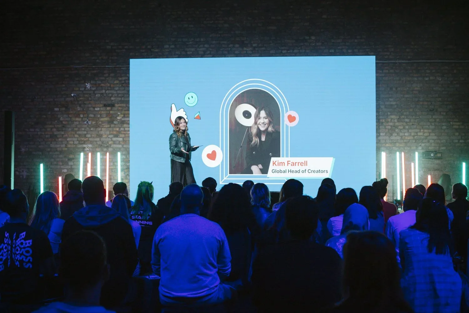 A woman stands on stage next to a large screen displaying a photo of Kim Farrell, labeled as Global Head of Creators. The audience watches the presentation in a dimly lit, industrial-style venue with exposed brick walls and colorful vertical lights.
