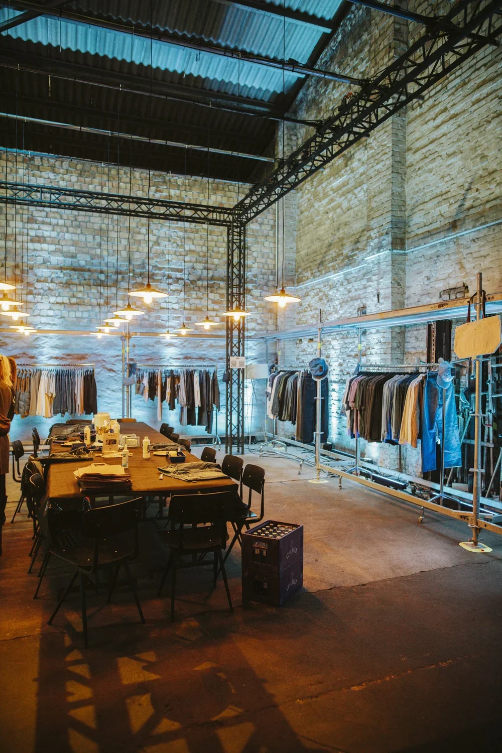 Clothing store or showroom with racks of hanging garments against brick walls, a large table with clothes and bottles, and industrial lighting in a spacious warehouse-like setting.