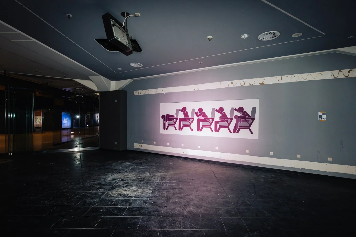 Empty room with a mural of stylized figures sitting at desks, taken in a dimly lit building with black tile flooring and a dark ceiling.