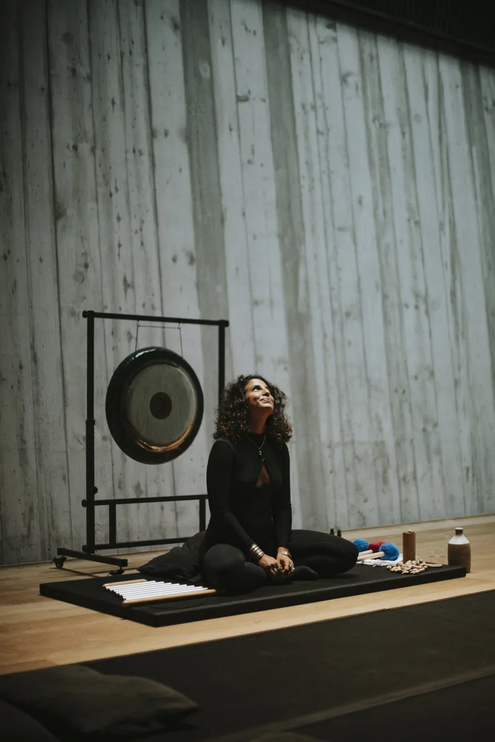 A woman with curly hair practicing sound healing with a gong in a minimalist studio with wooden floors and a concrete wall, sitting cross-legged on a mat surrounded by healing instruments.