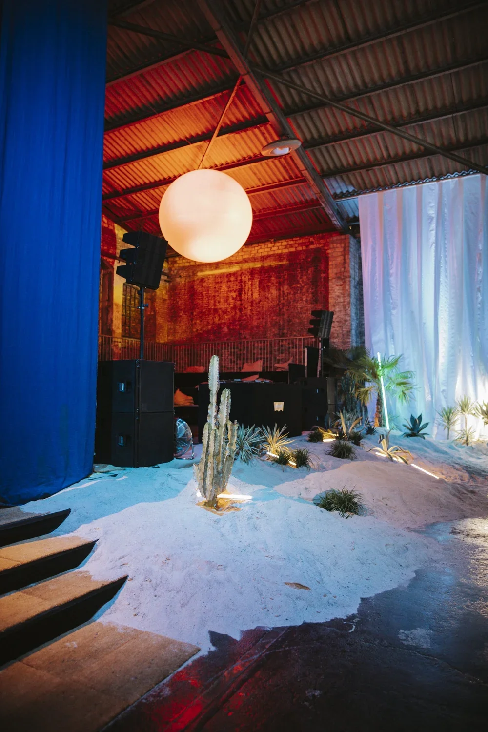 Indoor event space with sandy floor, desert plants, illuminated hanging lamp, and DJ equipment, set against a brick wall and white drapery with colorful lighting.