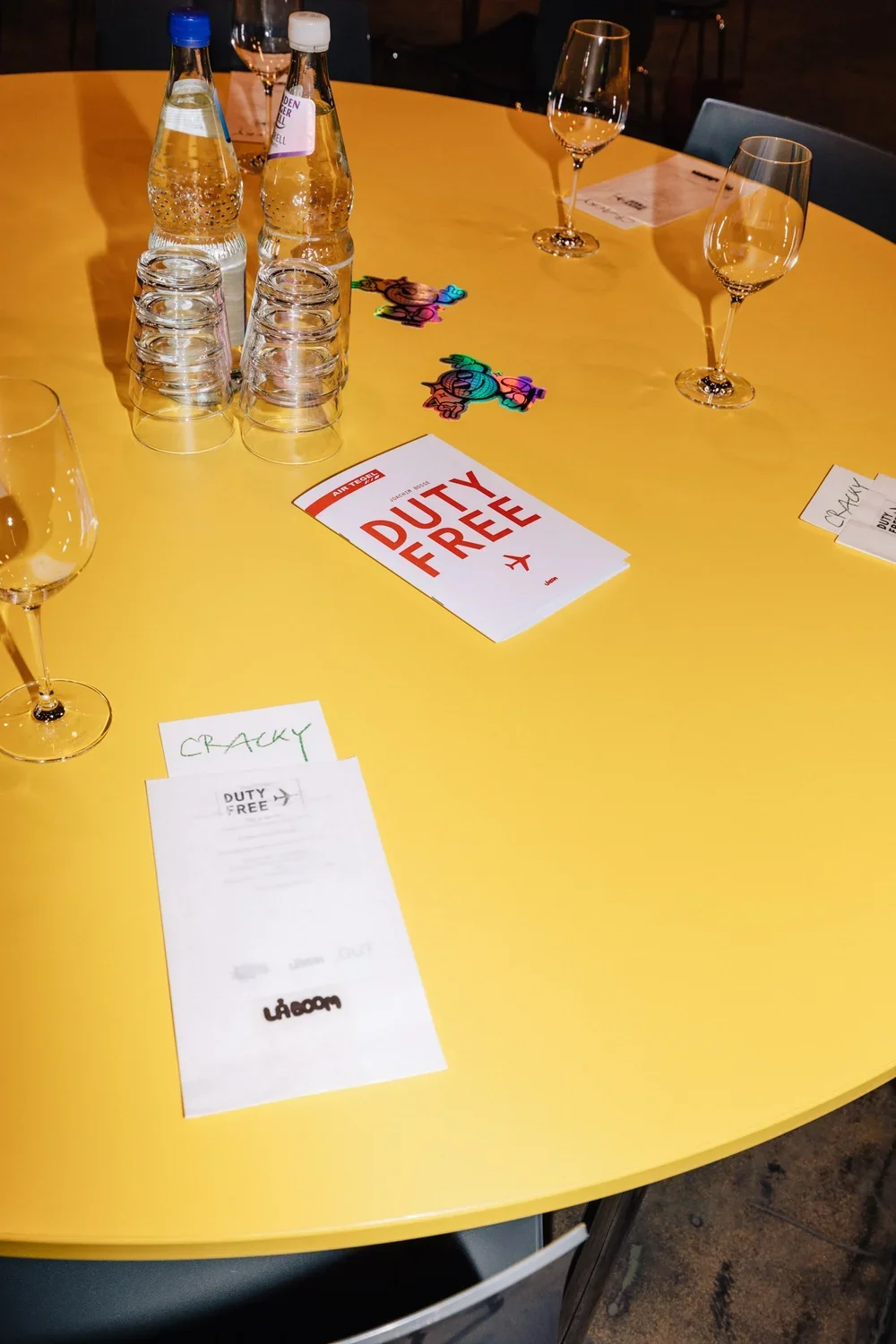 A round yellow table set for a duty-free shopping event, with bottled water, glasses, colorful rainbow stickers, and paper signs.