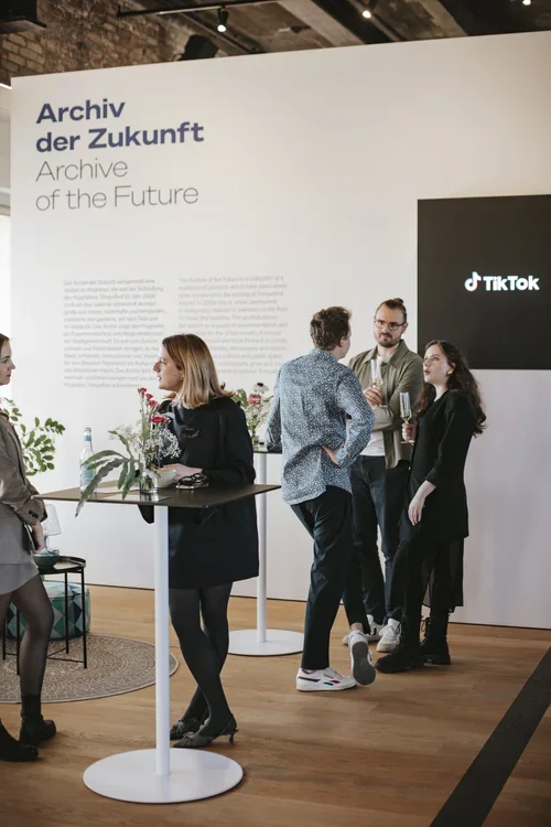 Group of five people engaging in conversation at an exhibition with signs reading "Archiv der Zukunft" and "Archive of the Future" in German and English, and a large TikTok logo on a screen.