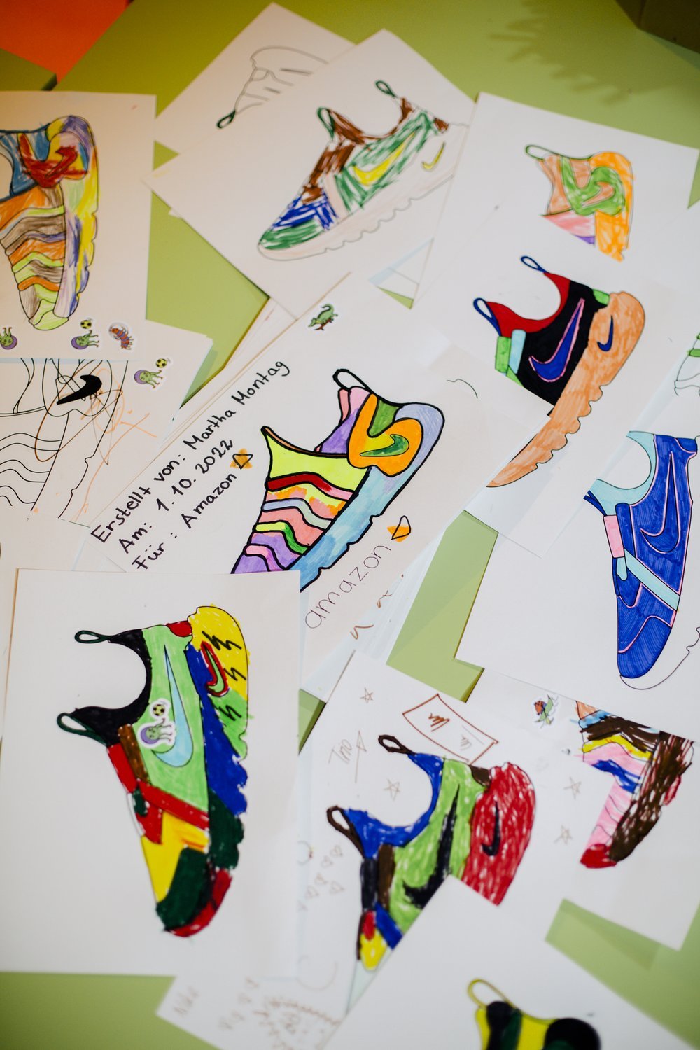 Multiple colorful drawings of sneakers on paper scattered on a green surface, with a handwritten note in German.