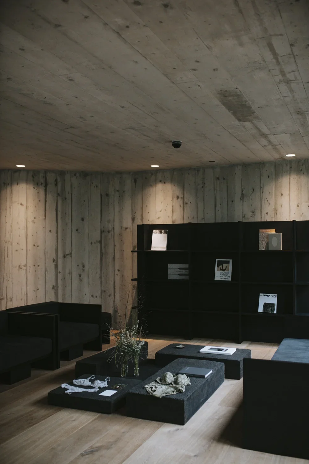 Modern minimalist living room with black furniture, a black bookcase, and a wooden wall and ceiling. The room has a wood floor with a small arrangement of textiles, books, and decor on a black low table, and a black armchair.