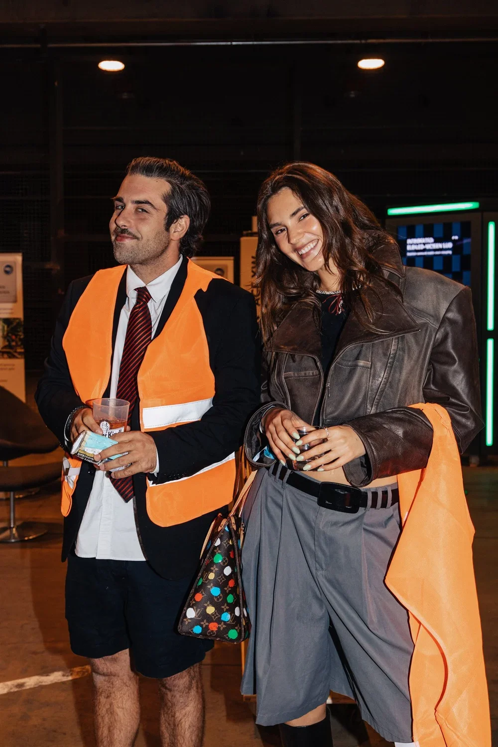 Two young adults smiling at the camera, one man and one woman, in an indoor setting. The man is dressed in a suit with a red striped tie and is wearing an orange safety vest, holding a drink and a credit card. The woman is wearing a leather jacket, g